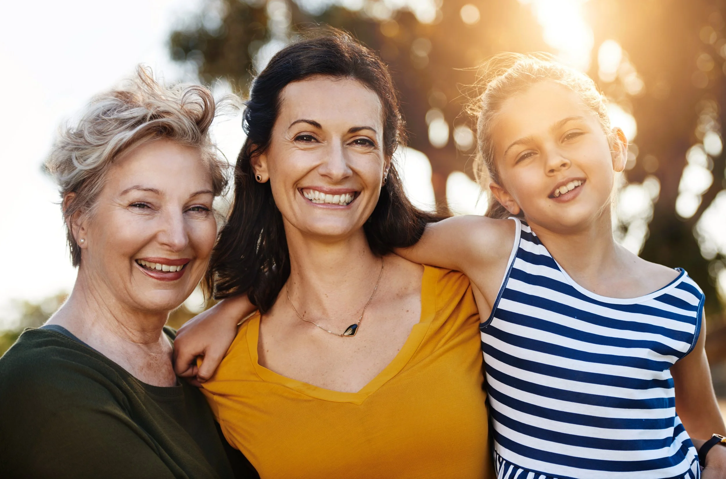Three females smiling outdoors with sunlight in the background, embracing each other. The older woman has short gray hair, the middle woman has long dark hair, and the young girl has long blonde hair in a striped shirt.