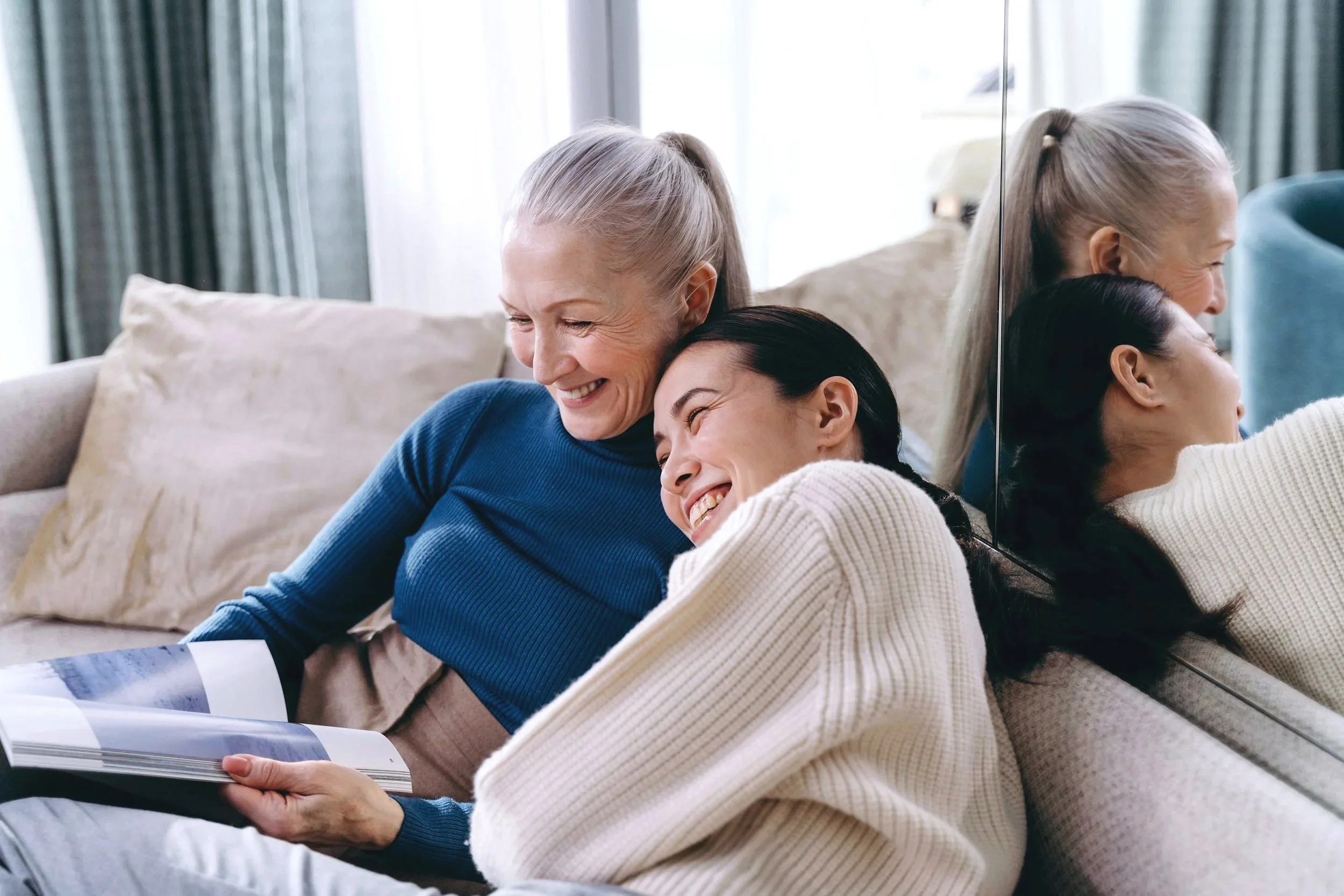 Two women, one older and one younger, sitting on a beige couch, smiling and leaning close to each other while looking at a photo album.
