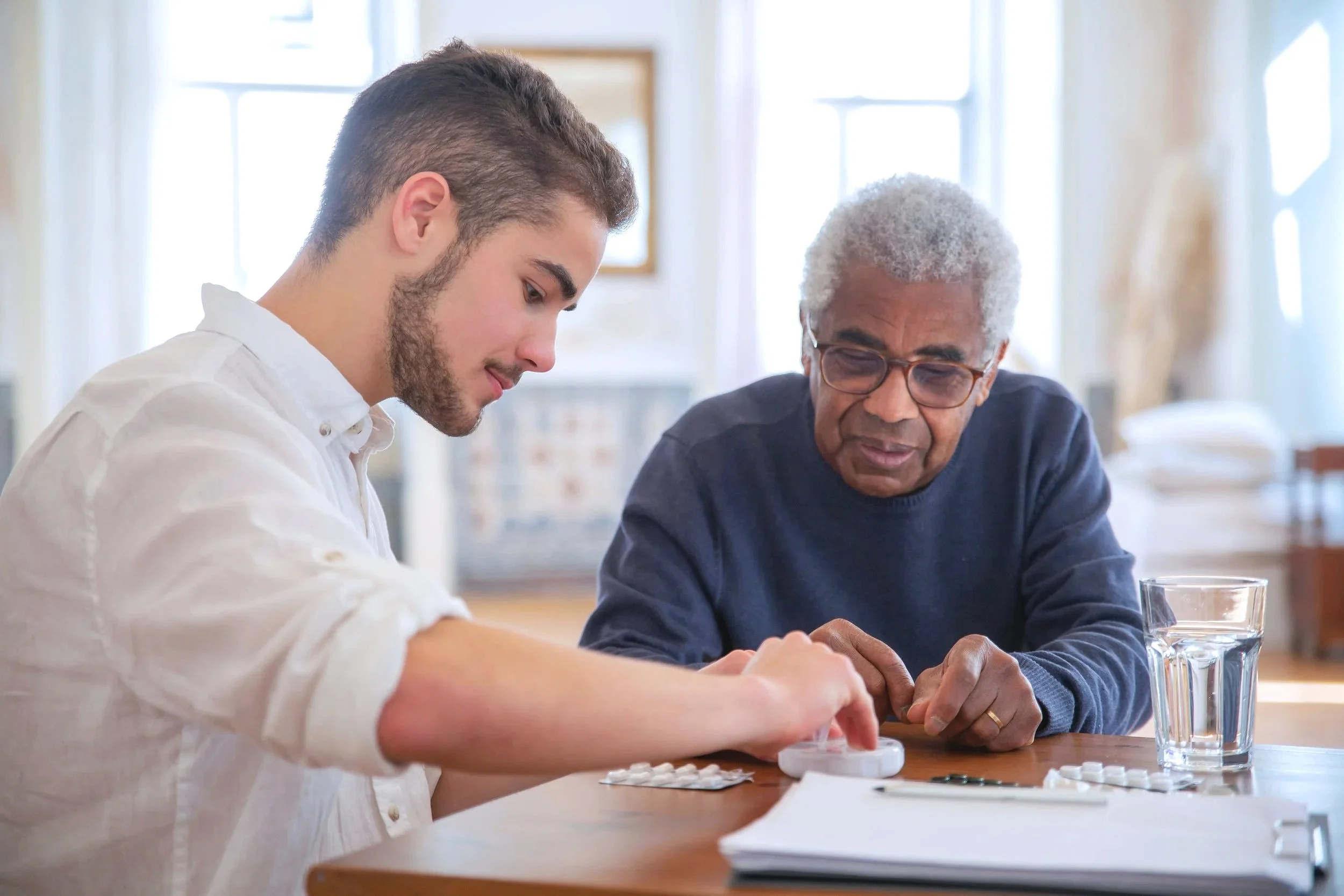 A young man and an elderly woman sitting at a table, looking at medication in a pill organizer.