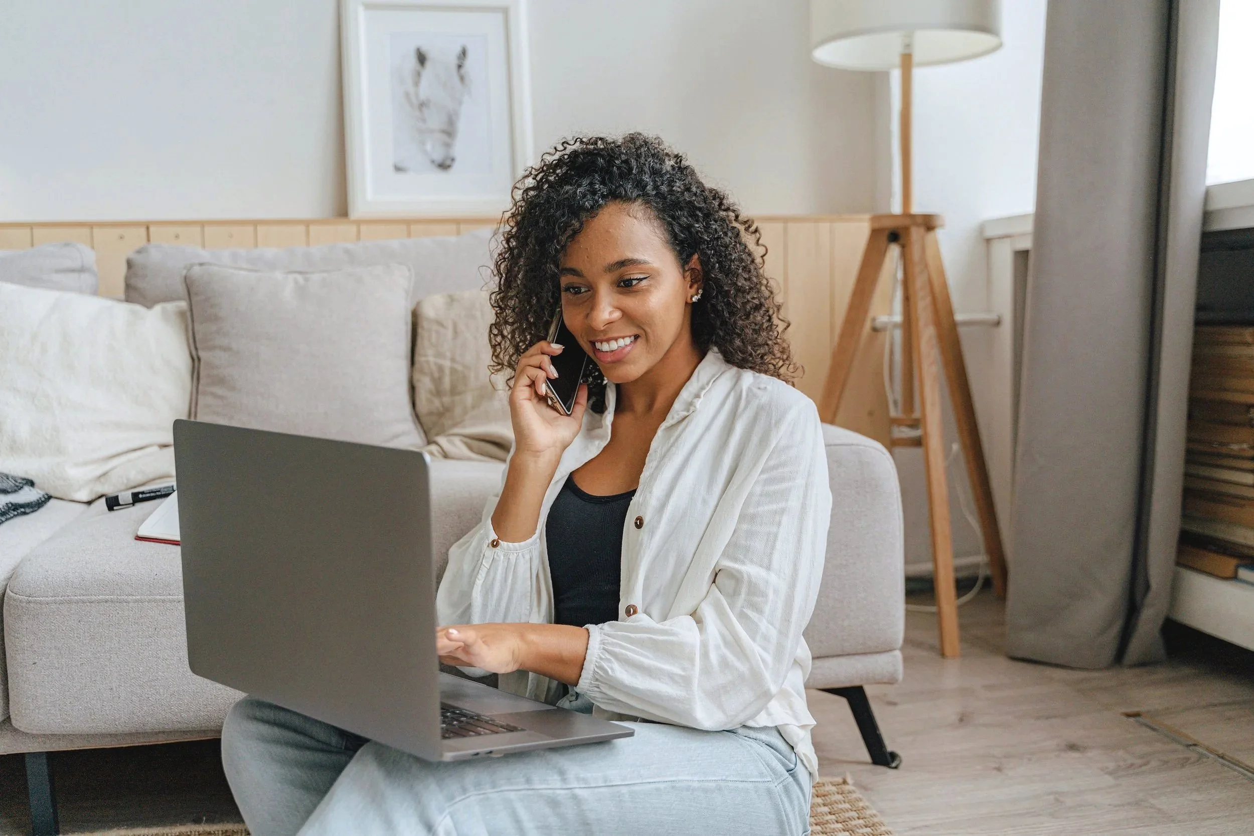 A woman with curly hair sitting on a couch, talking on her phone and working on a laptop in a cozy living room with beige and gray tones.