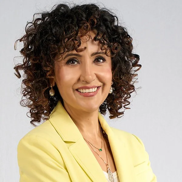 A smiling woman with curly dark hair wearing a yellow blazer and pearl earrings.