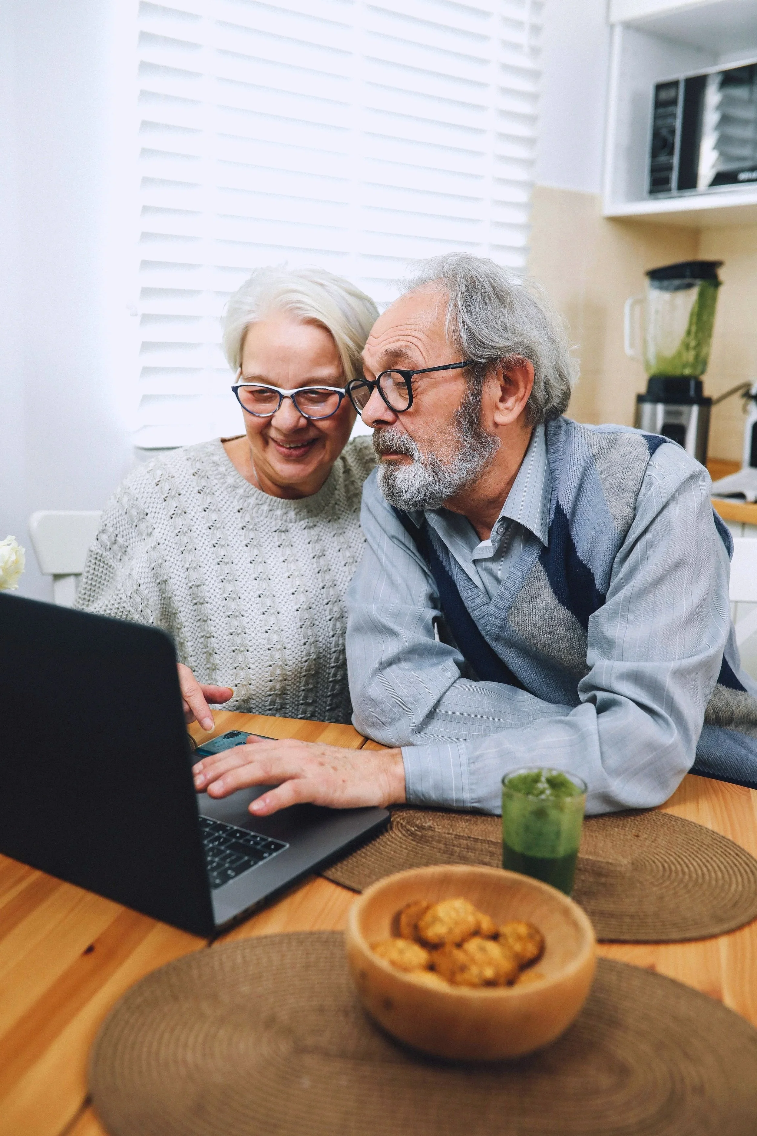 An elderly couple with glasses sitting at a wooden dining table, looking at a laptop together and smiling. The table has a bowl of cookies, a green smoothie, and brown placemats. In the background, kitchen appliances are visible.