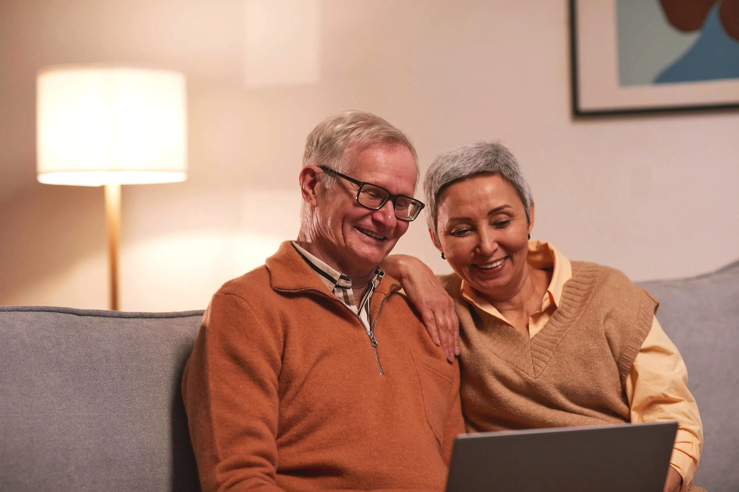 An elderly man and woman sitting on a gray couch, looking at a laptop together and smiling warmly in a cozy living room with a lamp and framed artwork in the background.