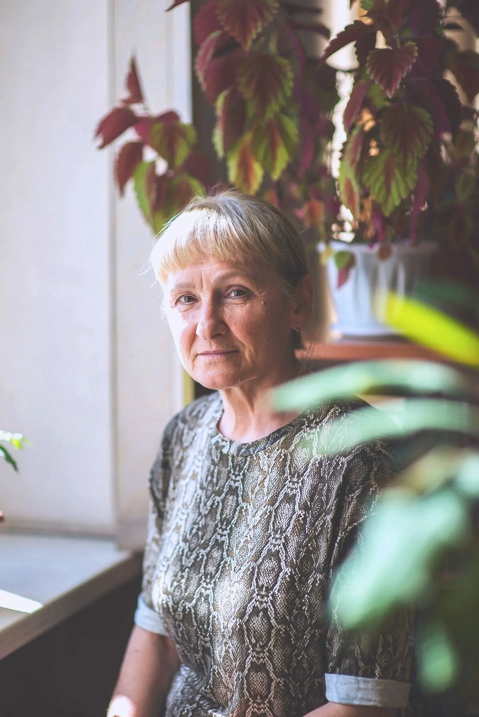 An elderly woman with short blond hair sitting near a window surrounded by green and reddish leaves, wearing a snake print shirt, looking at the camera.