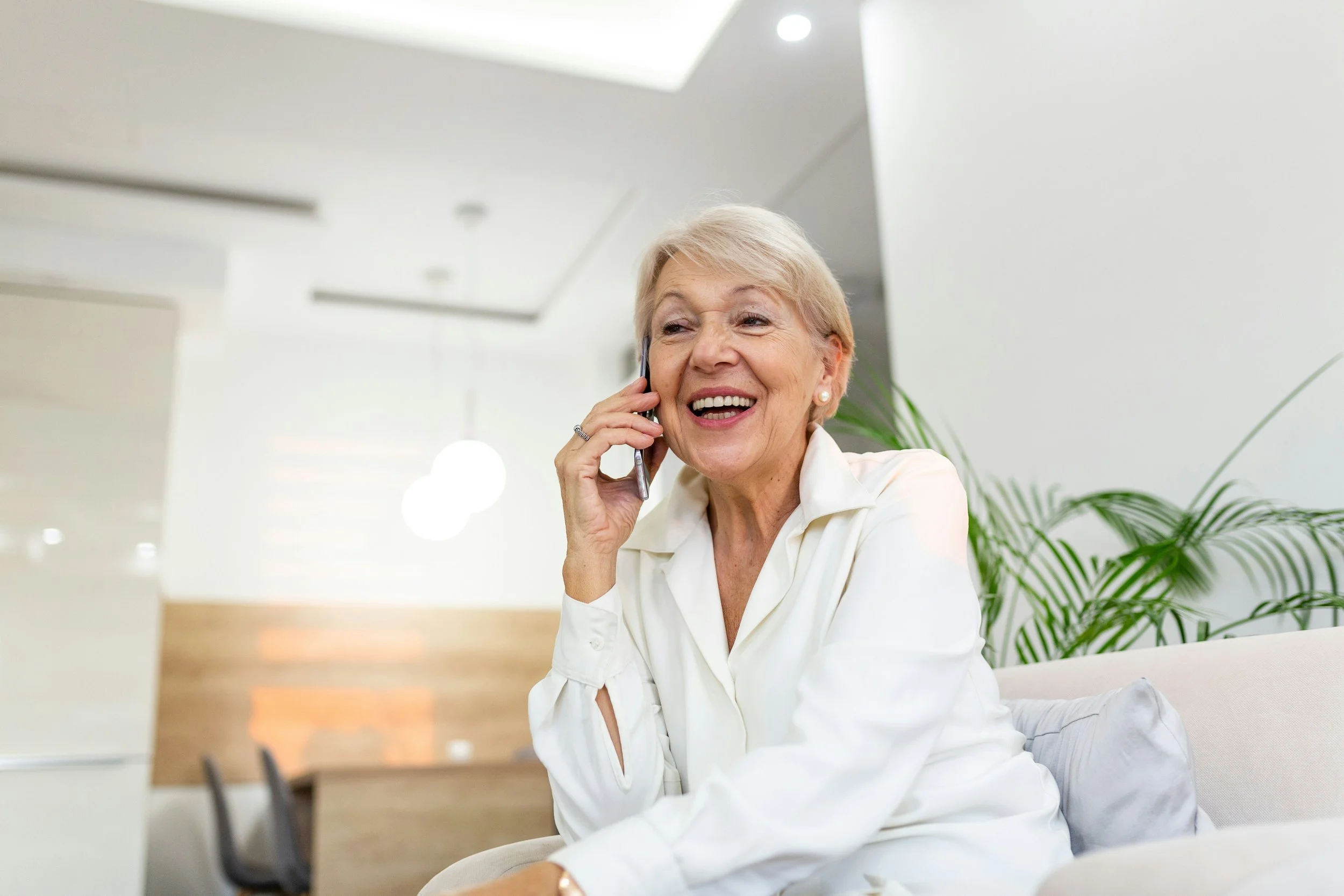An elderly woman with blonde hair, smiling and talking on her cellphone, sitting on a light-colored sofa in a modern, well-lit room with green plants.