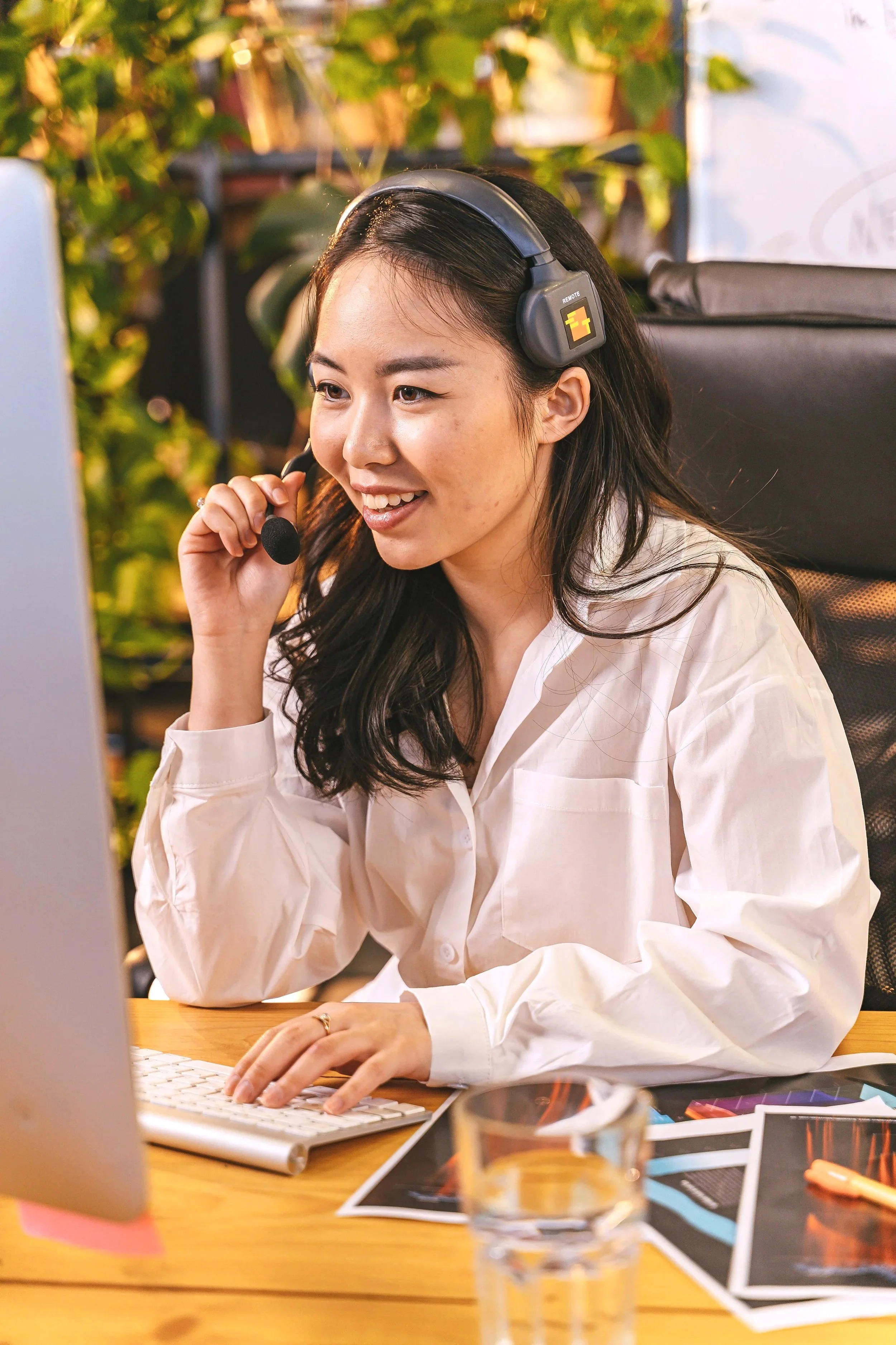 A woman wearing a white shirt and headset sitting at a desk with a computer, papers, and a glass of water, smiling and speaking into a microphone.