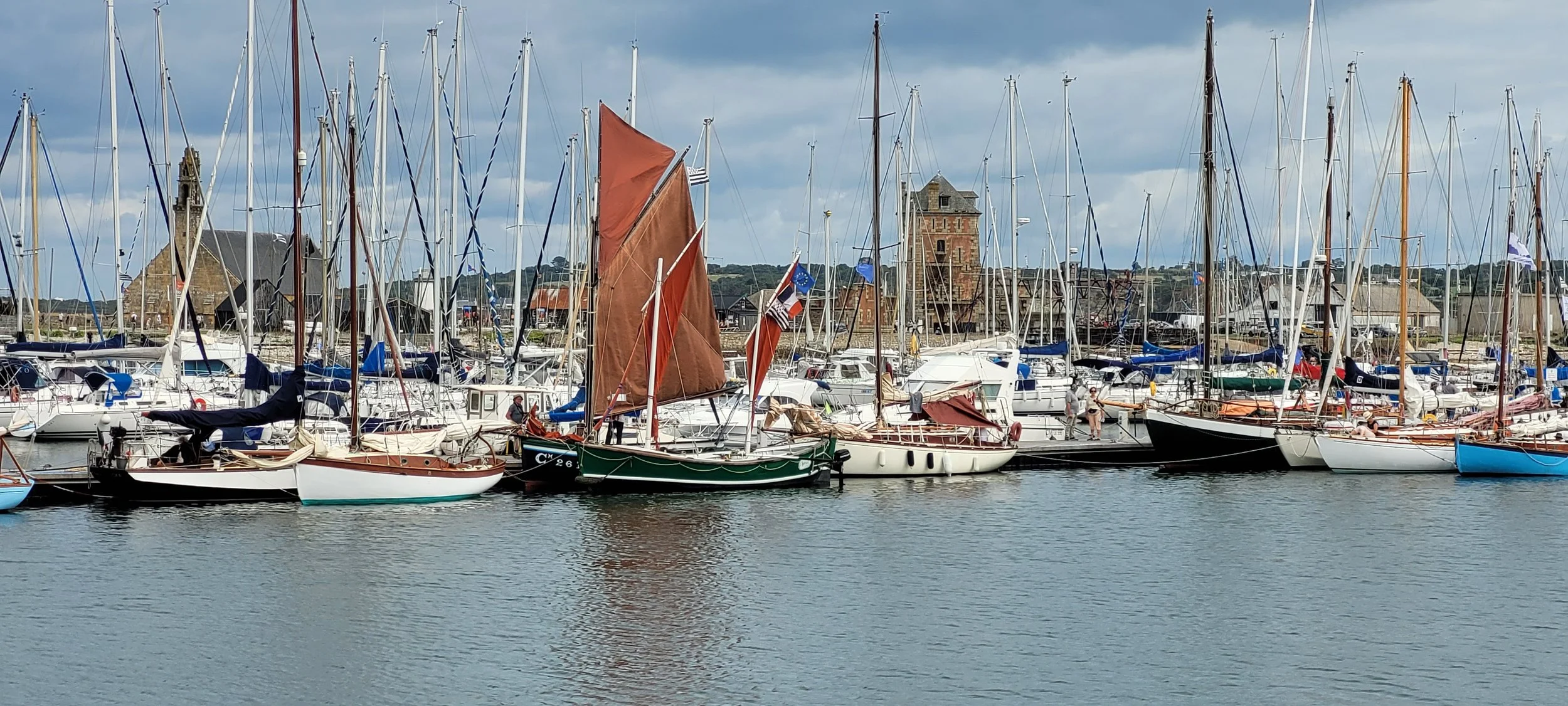Port de plaisance avec de nombreux voiliers amarrés, certains avec des voiles en toile de couleur marron, eau calme, bâtiments historiques et ciel nuageux en arrière-plan.