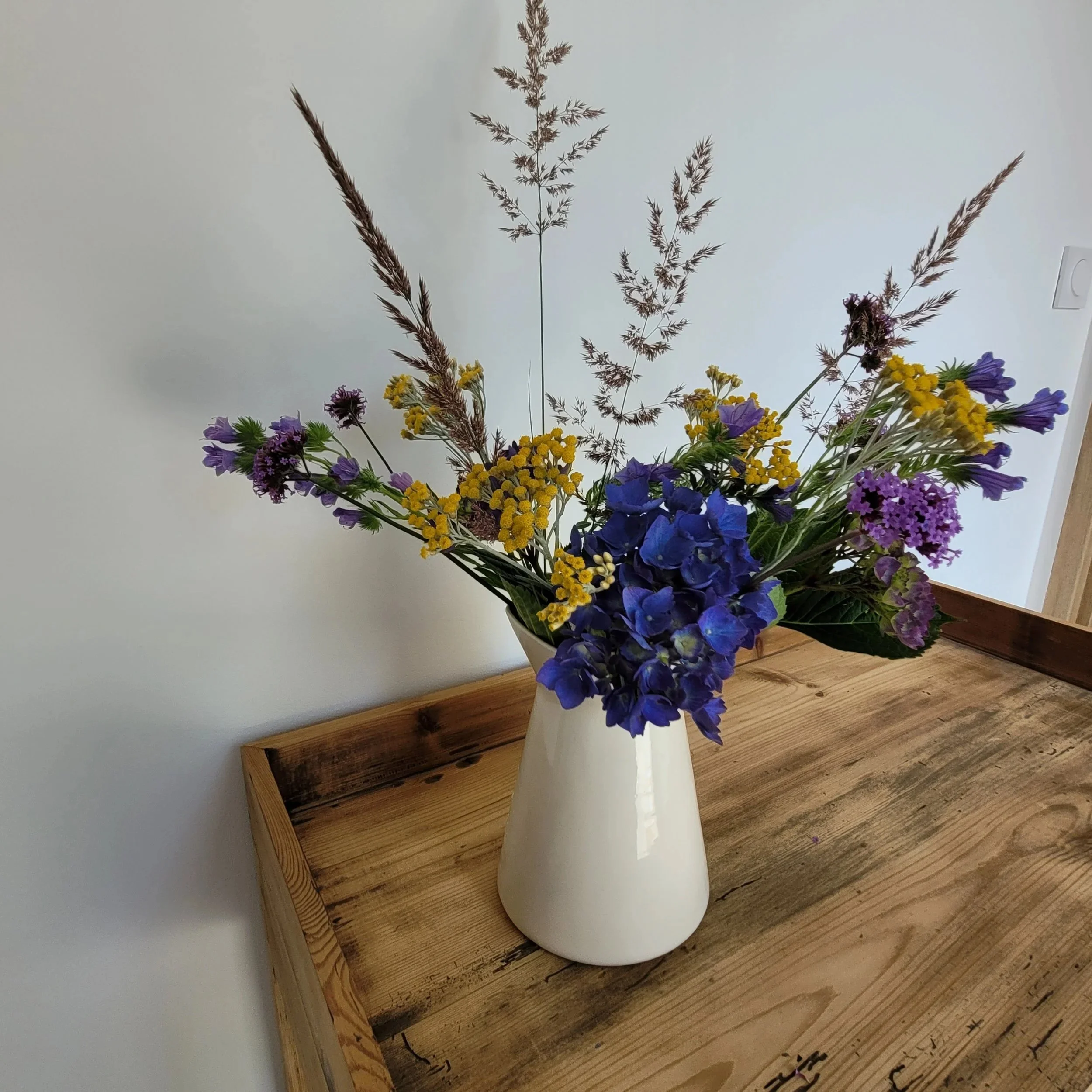 Un bouquet de fleurs colorées dans un vase blanc placé sur une table en bois, avec un mur blanc en arrière-plan.