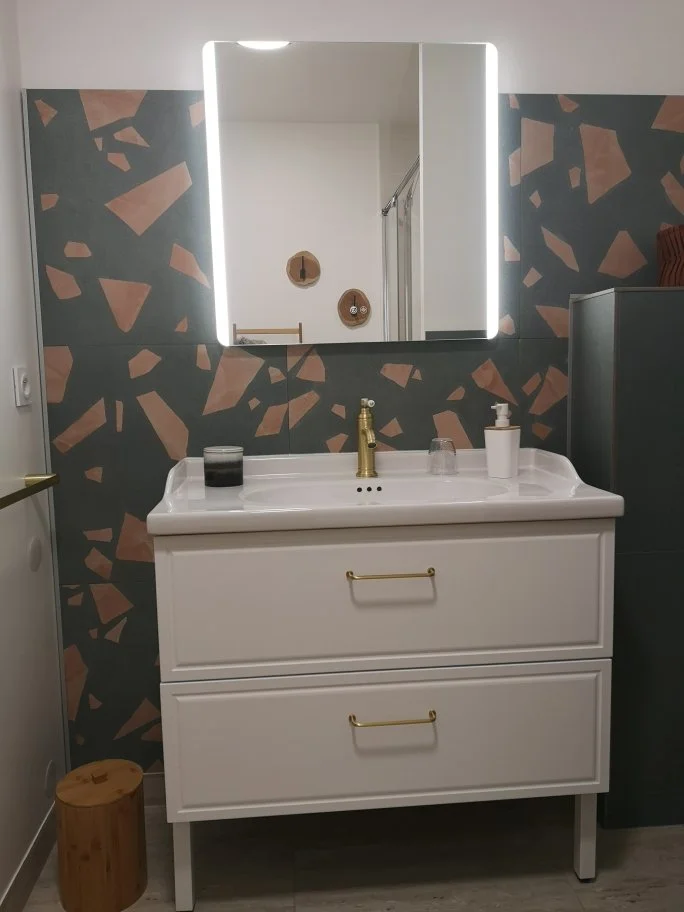 Un miroir éclairé au-dessus d'un lavabo blanc avec deux tiroirs, une poignée dorée, un savon et un verre d'eau, dans une salle de bain moderne avec un mur à motifs et des décorations murales.