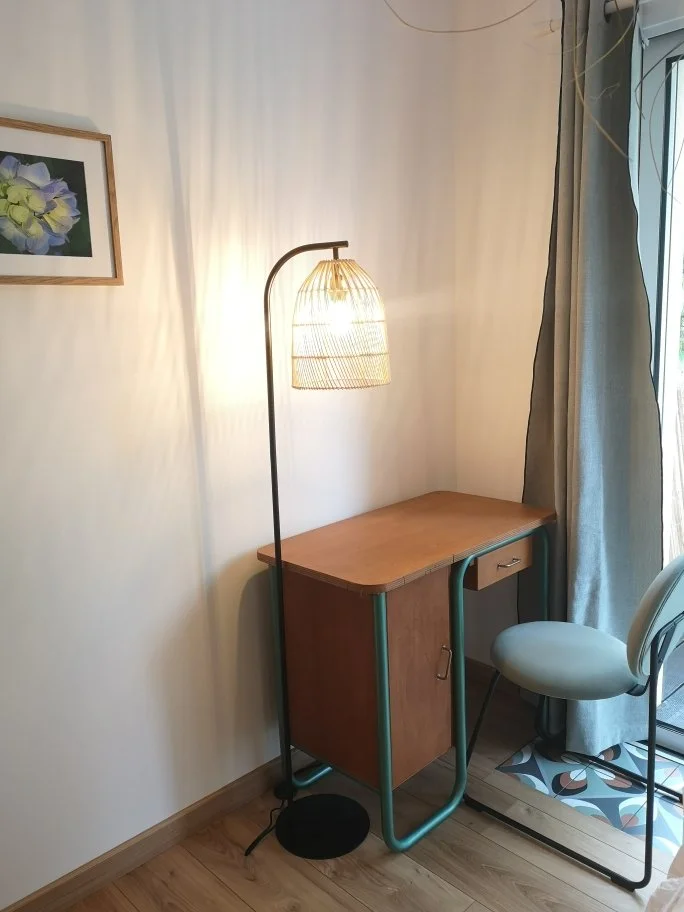 Une lampe de lecture sur un bureau en bois dans un coin de pièce avec une chaise beige, une fenêtre avec rideaux gris et un cadre avec une peinture de fleurs sur le mur.