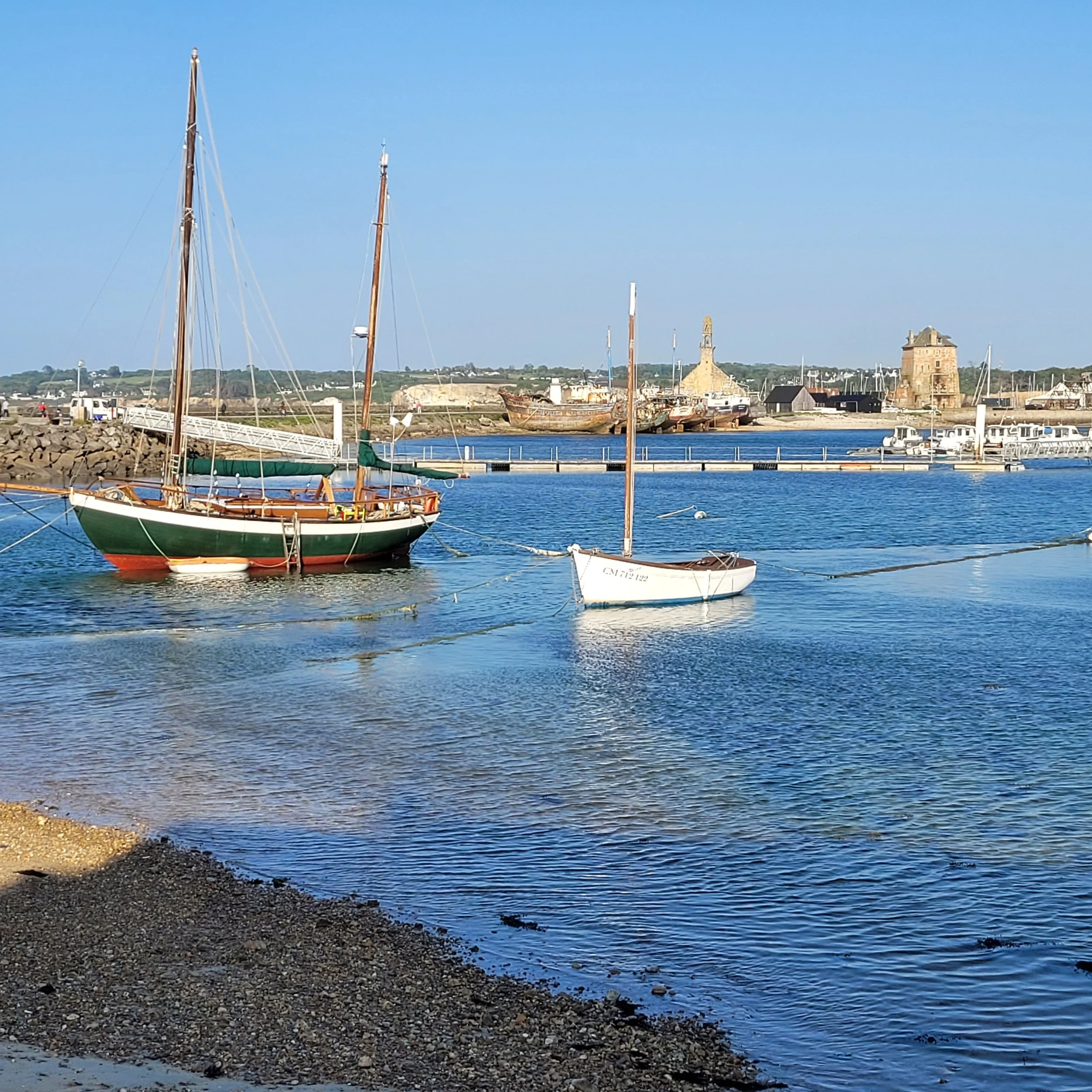 Un port de plaisance avec des bateaux à voile amarrés, dans un paysage maritime avec un ciel clair, un bâtiment historique en pierre en arrière-plan, et une plage de galets au premier plan.