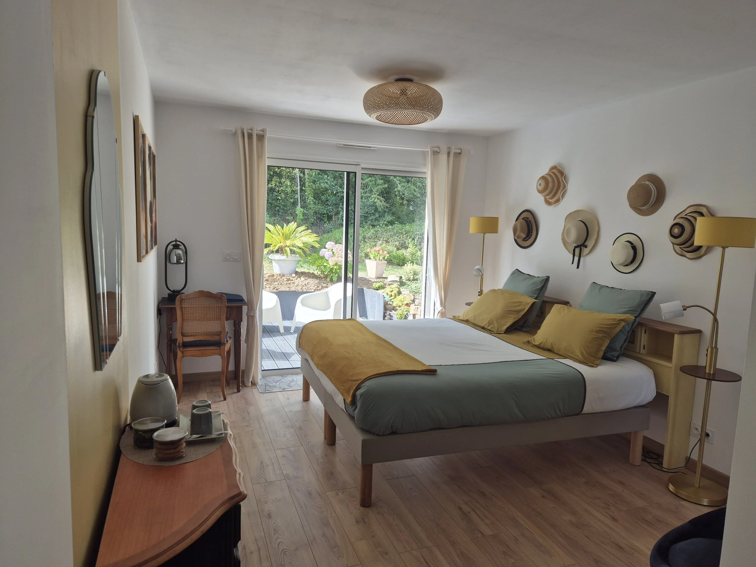 Chambre avec lit fait, volets ouverts sur un jardin avec des plantes et fleurs, décorée avec des chapeaux sur le mur, deux lampes de chaque côté du lit, grande fenêtre en verre.