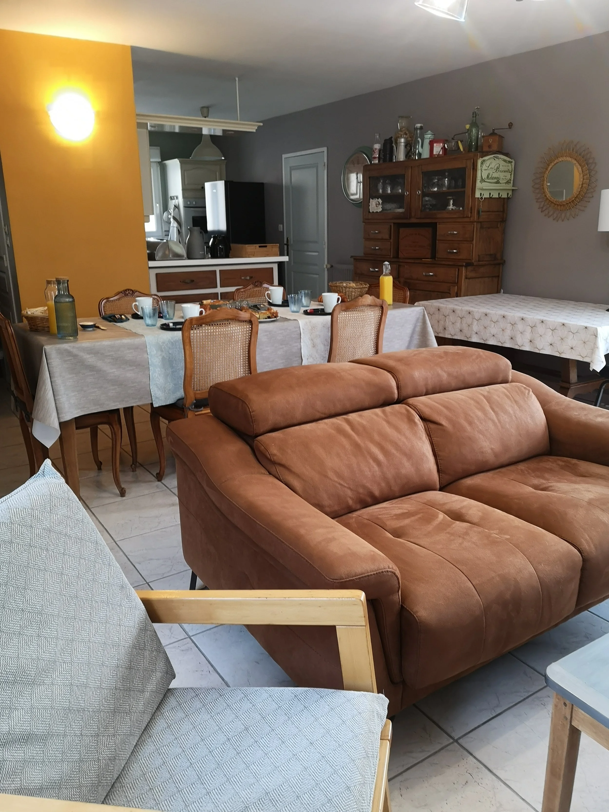 Une salle à manger avec une table décorée, plusieurs chaises en bois et une étagère en bois contenant des bouteilles et des objets décoratifs. Un canapé marron est visible au premier plan.