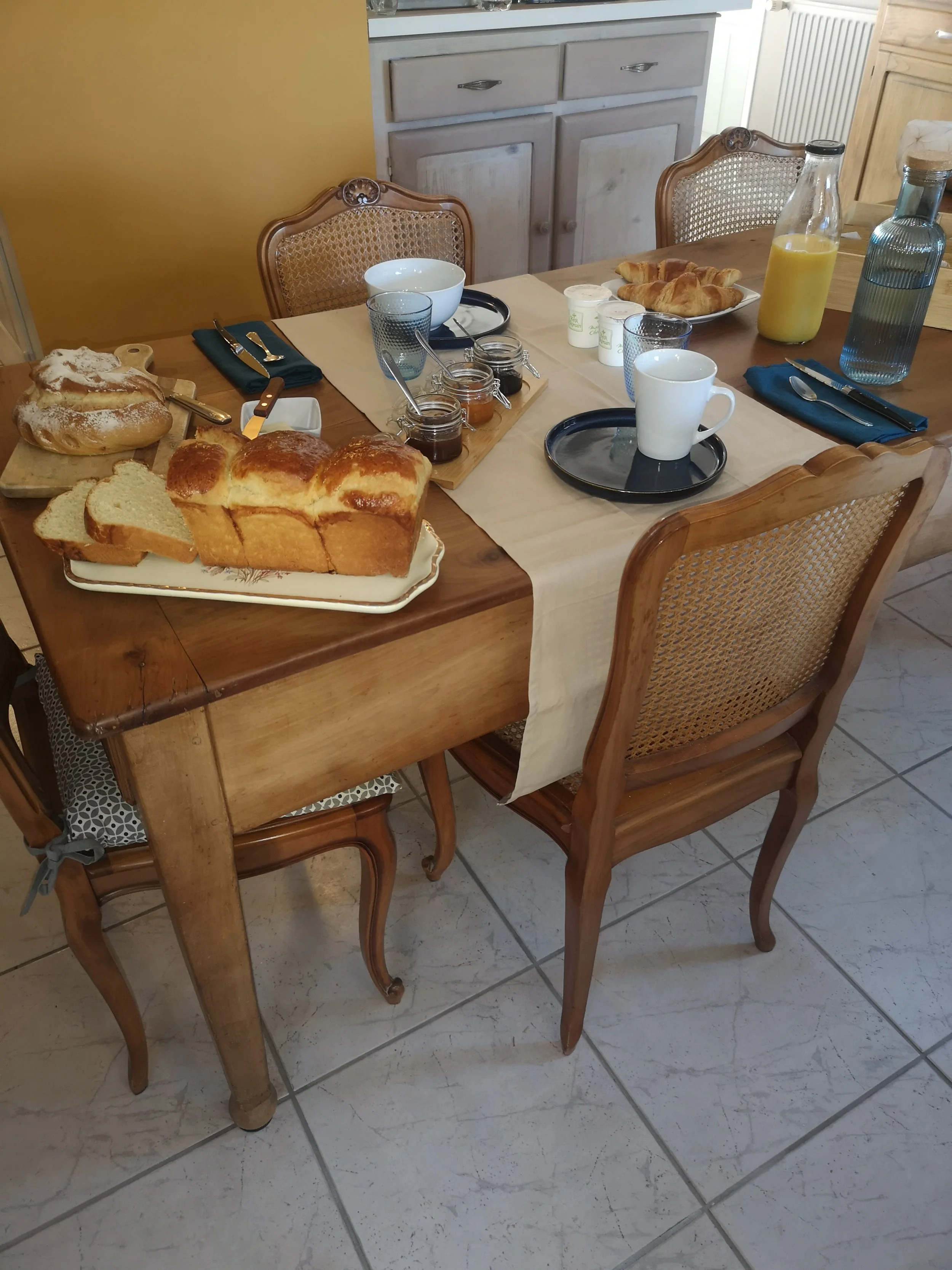 Table de petit déjeuner avec pain, confitures, croissants, jus d'orange, tasses, verres et assiettes, dans une cuisine rustique avec chaises en bois.