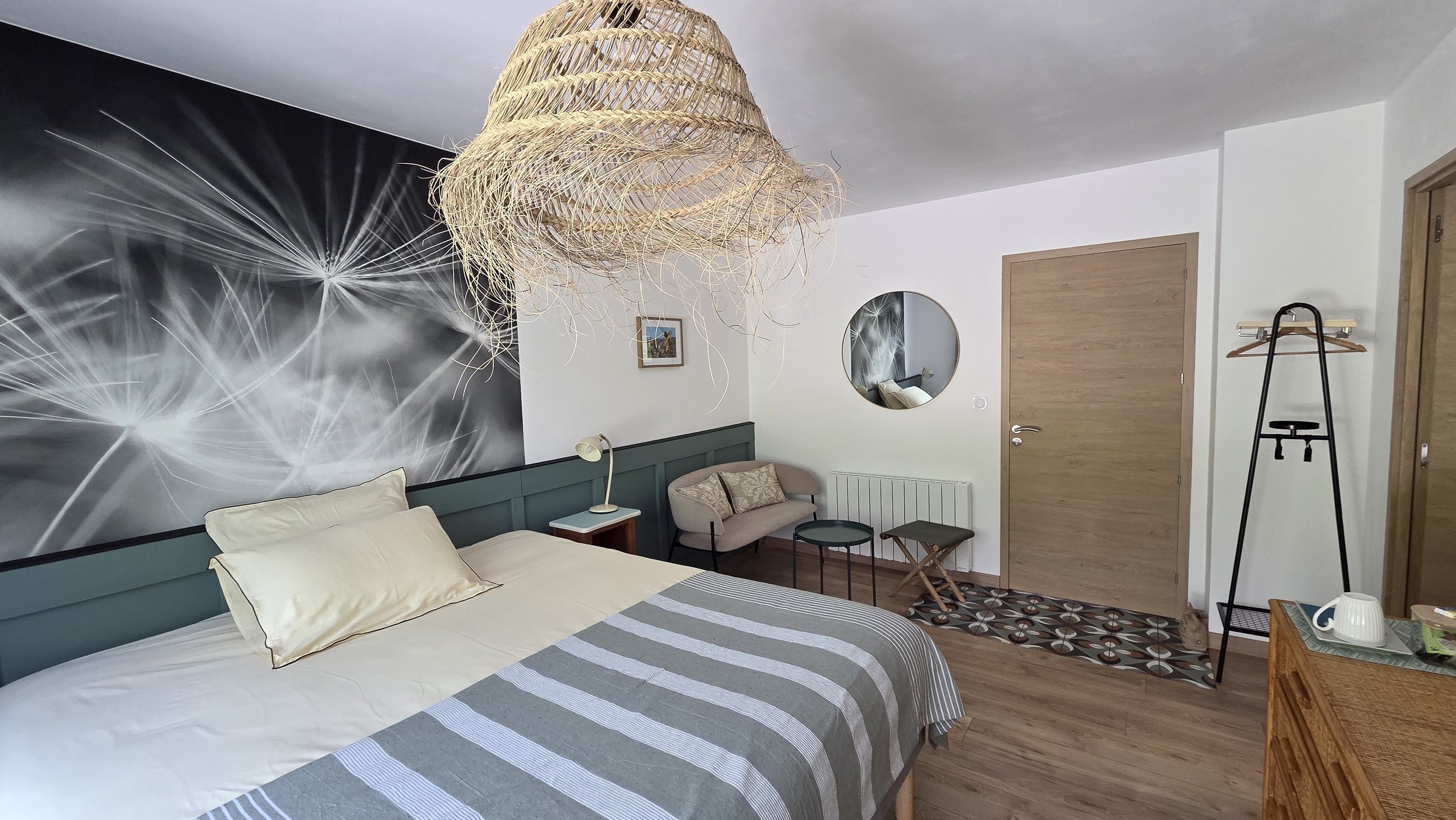 Chambre moderne avec délimitation murale représentant un motif d'éclats lumineux, suspension en rotin au plafond, lit avec linge rayé, fauteuils, miroir rond, porte en bois, et décorations variées.