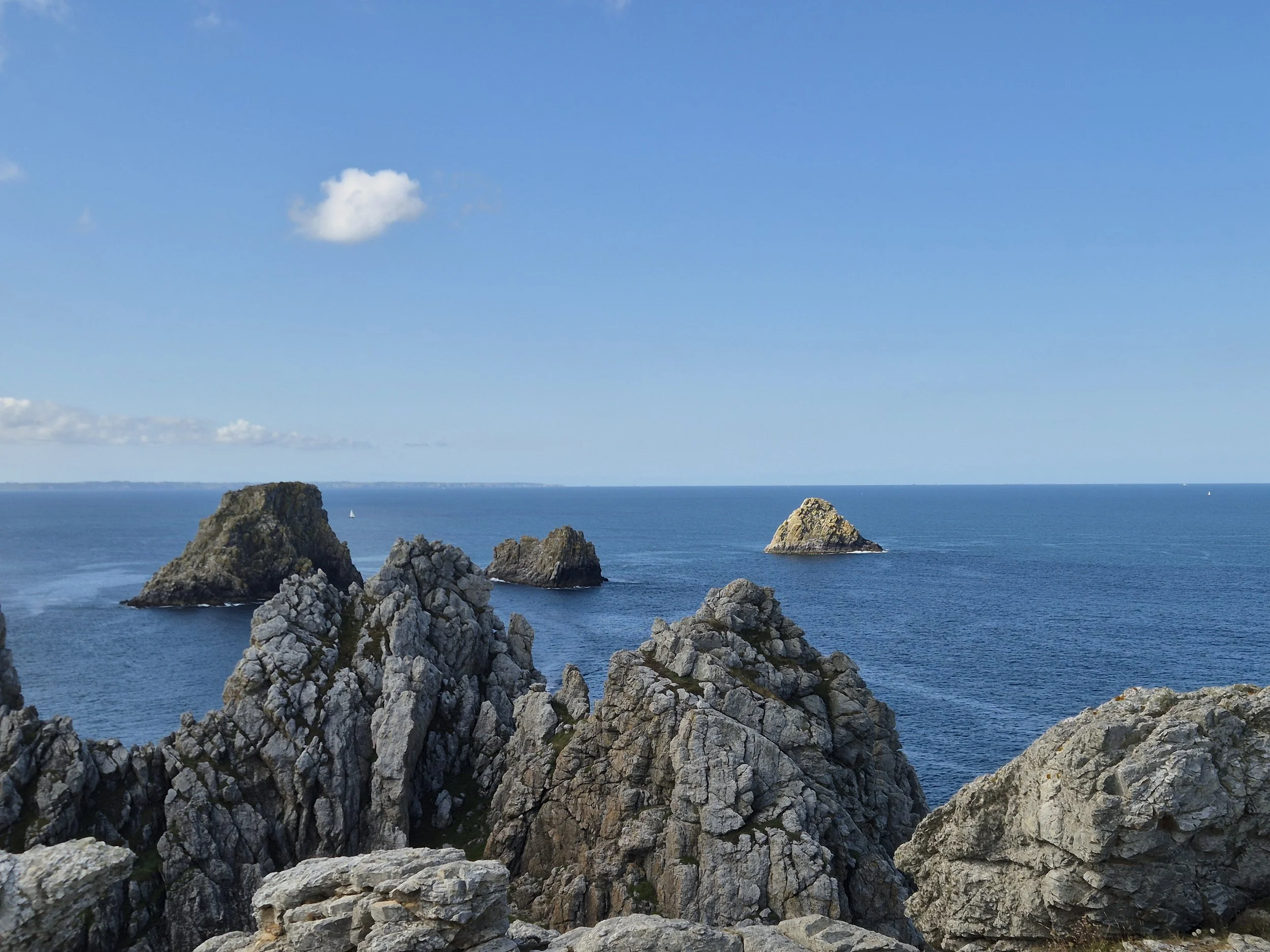 Ciel bleu avec quelques nuages, vagues de la mer, rochers pointus au premier plan qui encadrent la vue sur l'océan, petite île rocheuse au fond à droite.