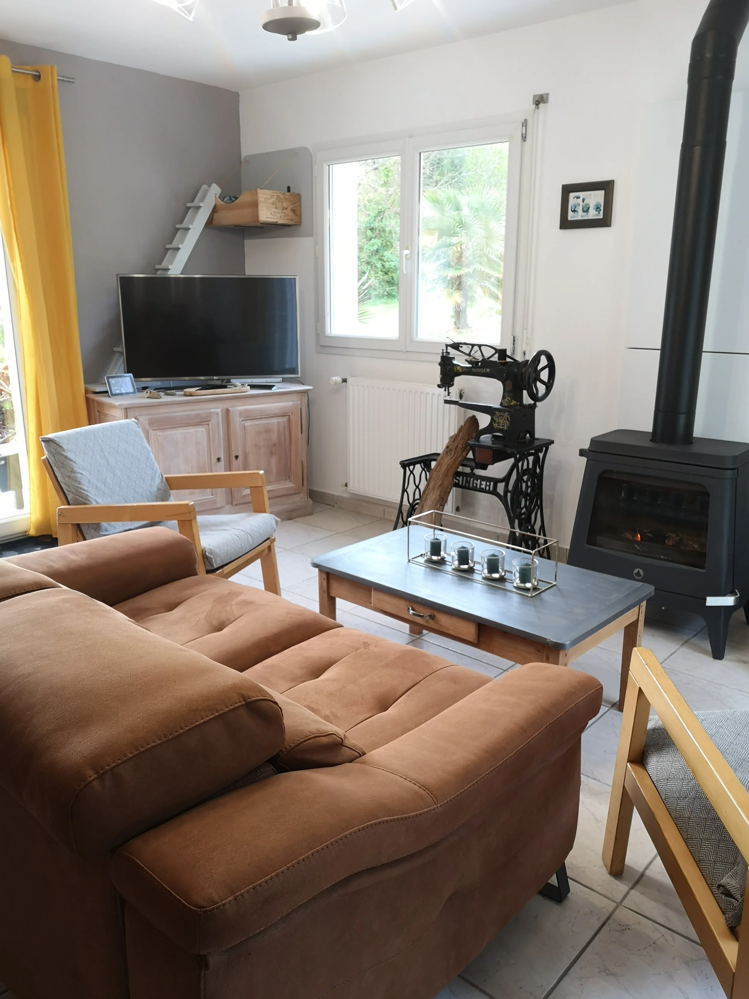 Vue d'un salon intérieur avec un canapé en cuir marron au premier plan, une table basse avec des bougies, une télévision sur un meuble en bois, une cheminée, une chaise en bois avec un coussin, une fenêtre laissant entrer la lumière, et un mur blanc avec une petite œuvre d'art.