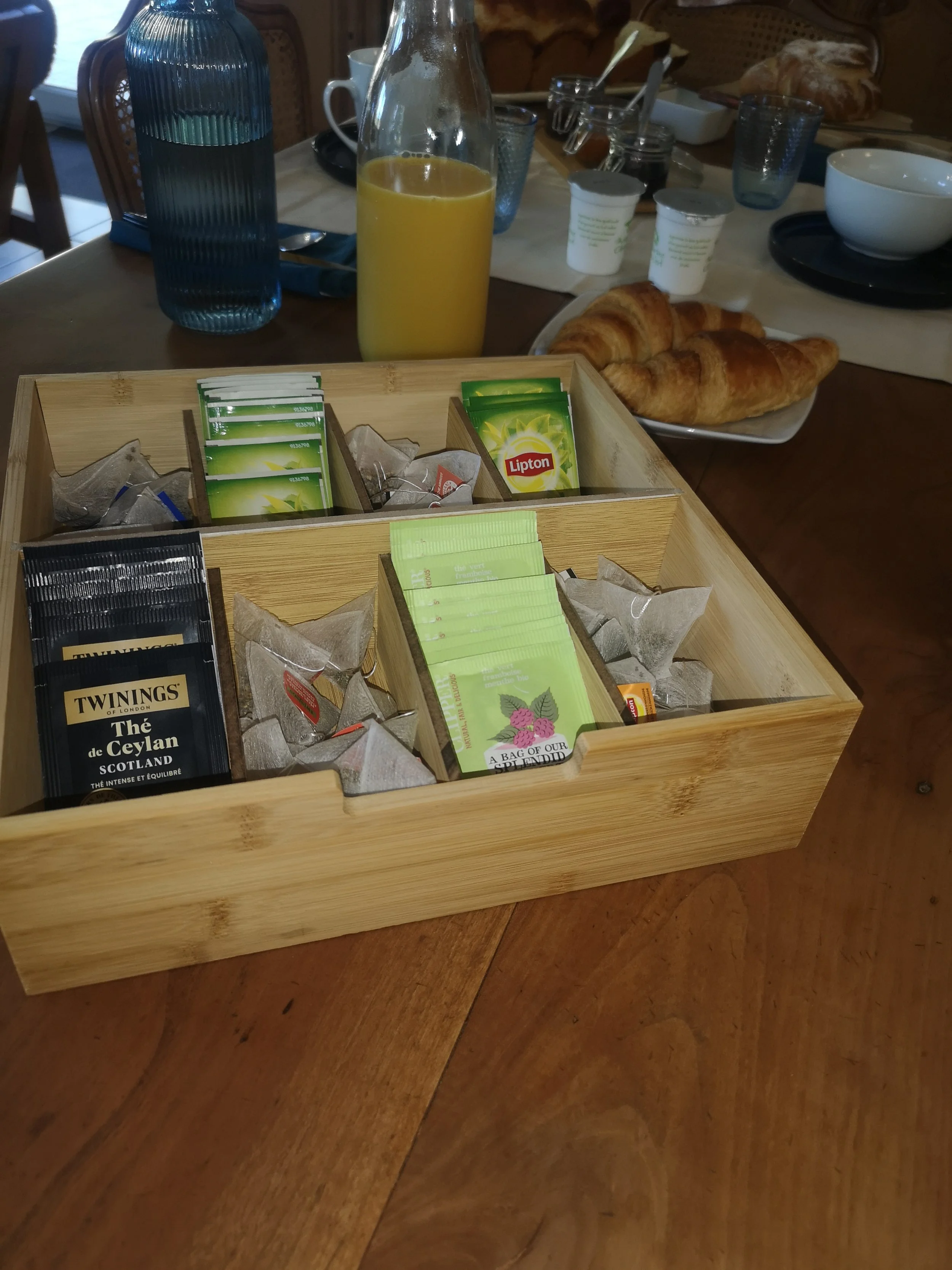 Un plateau en bois avec des sachets de thé, du sucre et des sachets de Lipton. En arrière-plan, un plateau avec deux croissants, une bouteille de jus d'orange, des tasses, des verres et des petits pots de confiture sur une table de petit déjeuner.