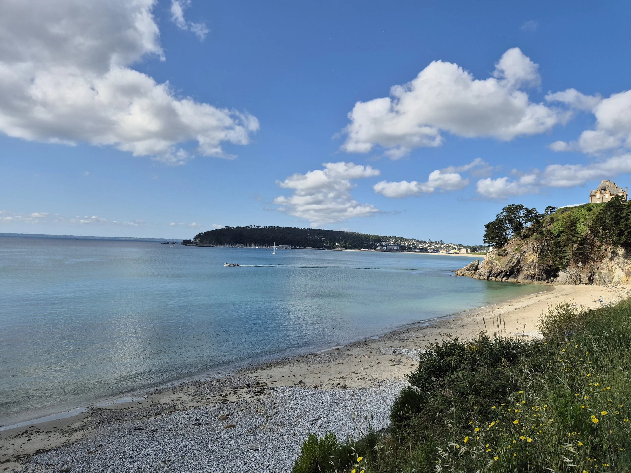 Plage paisible avec sable et galets, surplombée par une falaise avec une maison en pierre, vue sur la mer calme avec quelques bateaux, ciel bleu avec quelques nuages.