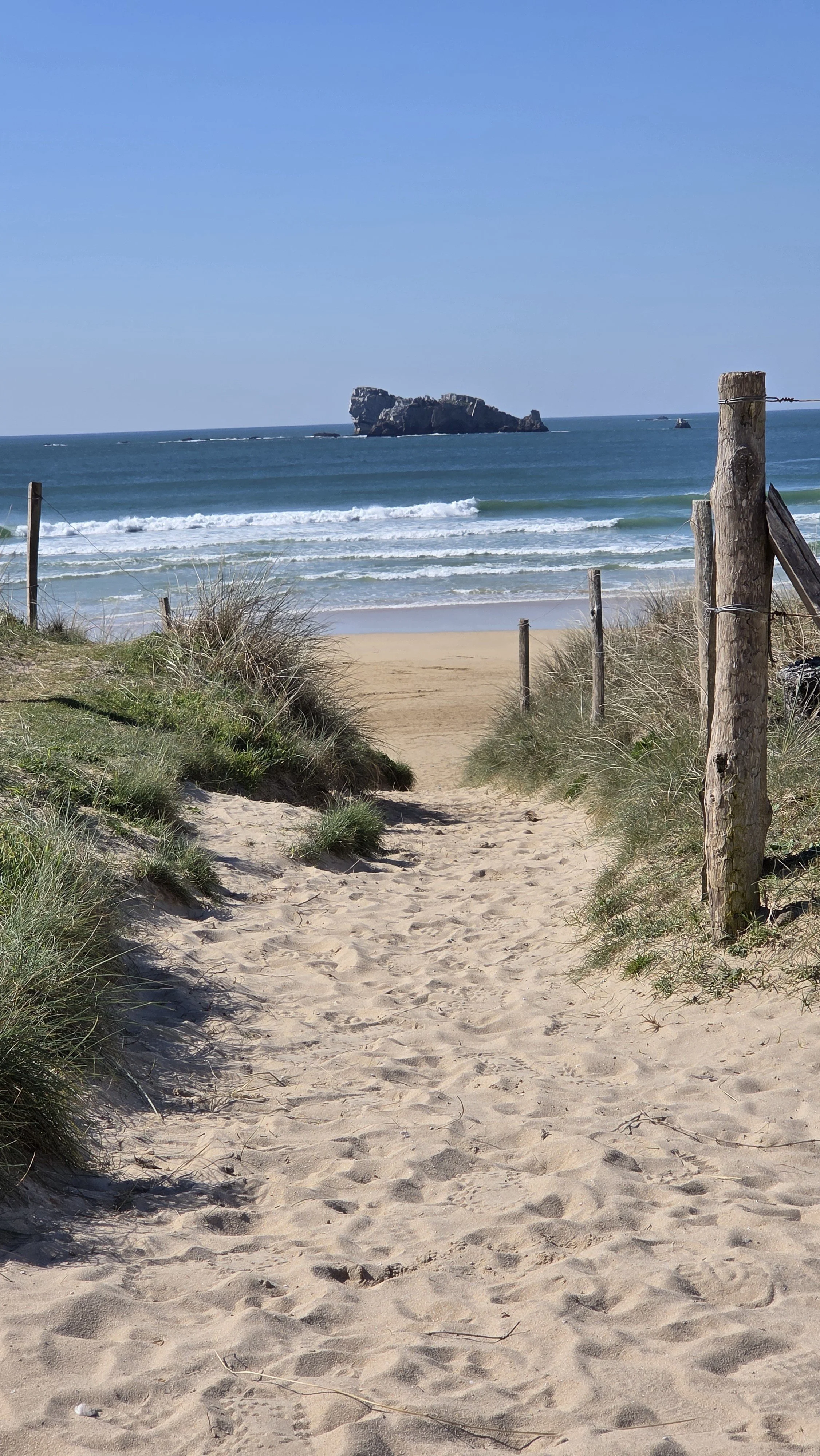 Sentier de sable menant à une plage avec vue sur la mer, rochers au loin, ciel clair, et dunes avec herbes hautes.