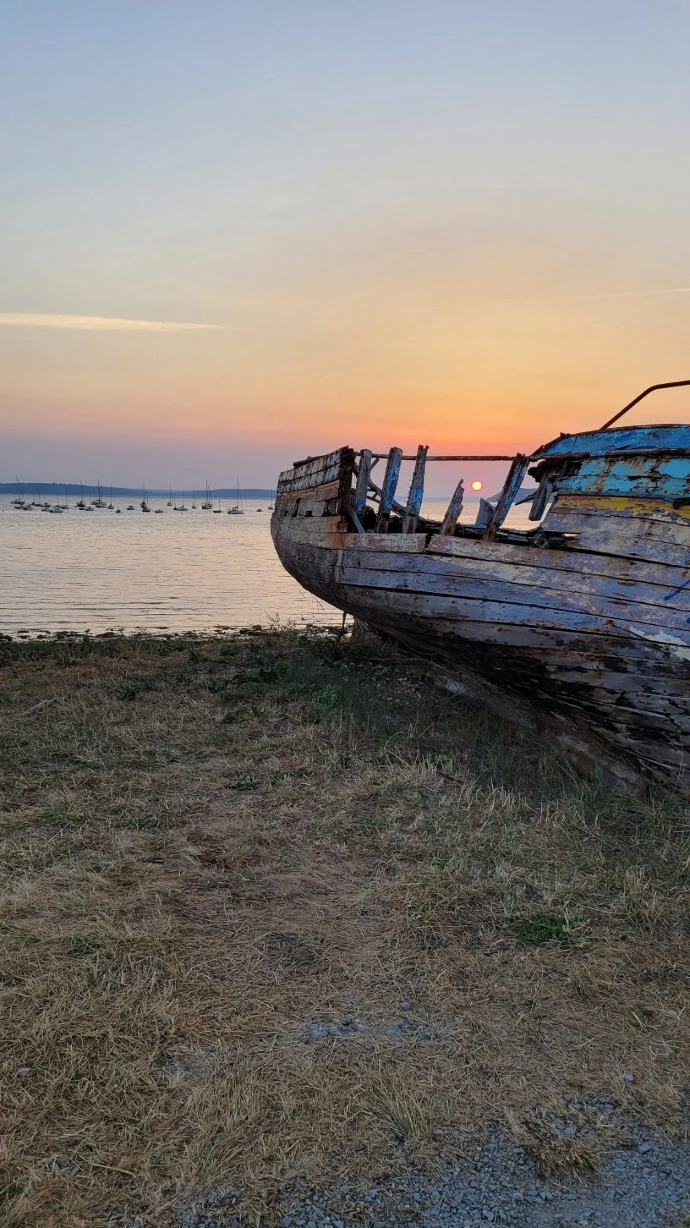 Un bateau en bois abandonné sur la rive, avec un coucher de soleil en fond et des voiliers en mer.