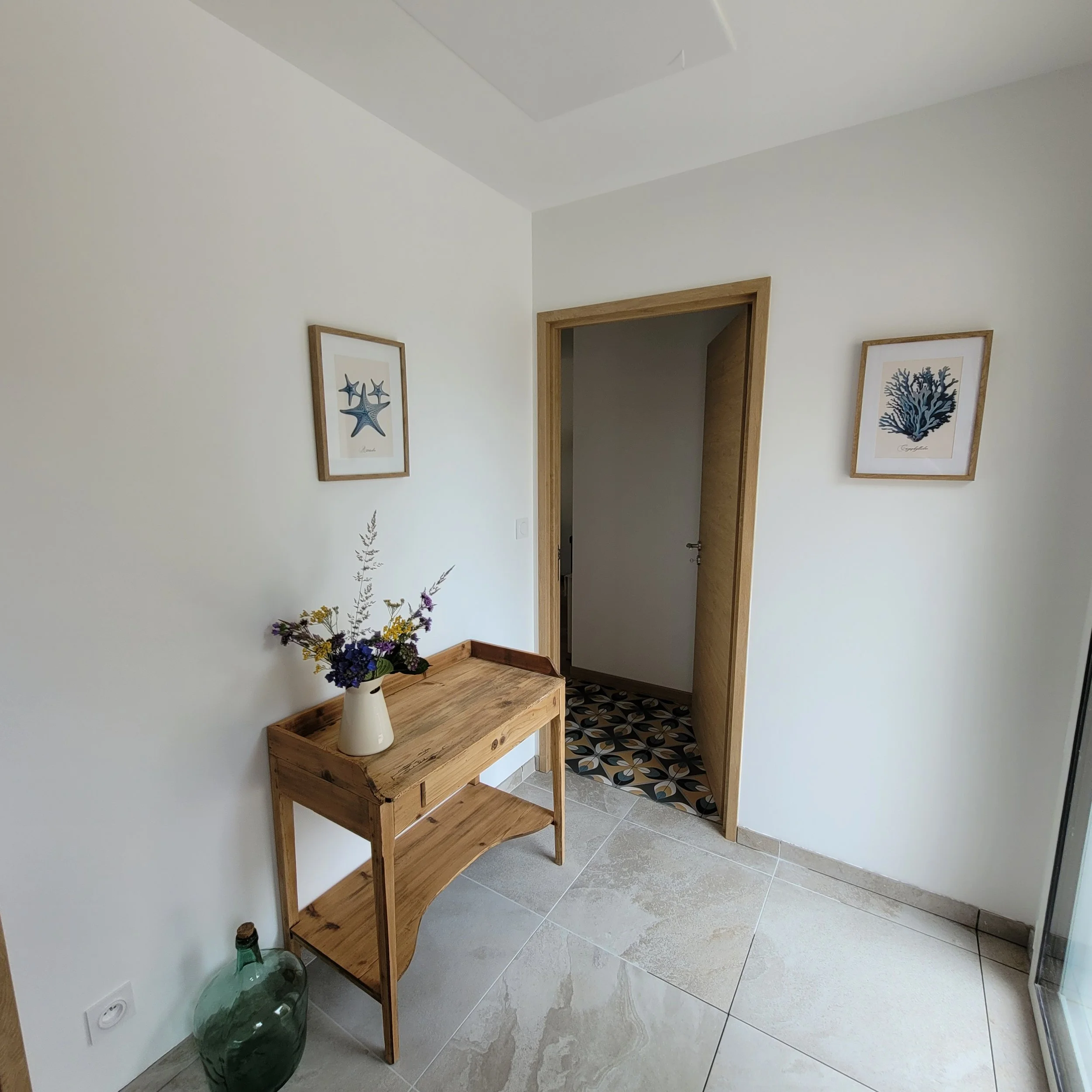 Un couloir ou un hall d'entrée avec des murs blancs, une porte en bois, et deux tableaux encadrés représentant des coraux ou des étoiles de mer. Il y a une table en bois avec un vase rempli de fleurs et une grande jarre en verre à côté. Le sol est en carreaux gris. La pièce est bien éclairée.