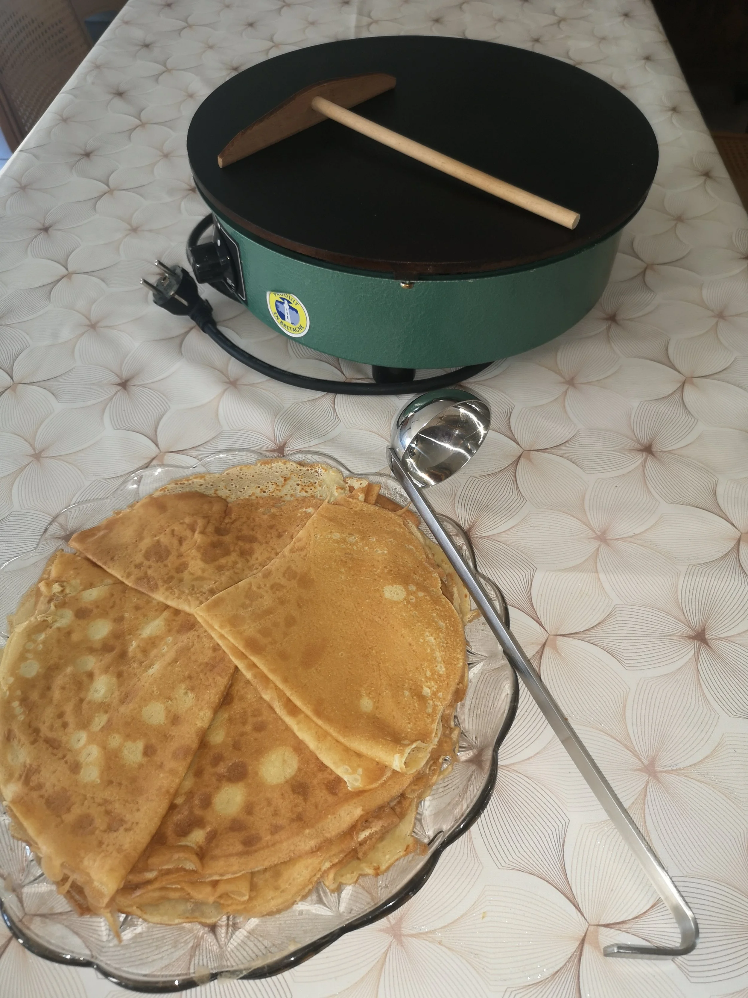 Une assiette de crêpes sur un plateau en verre, avec une cuillère et un appareil électrique de cuisson à crêpes en arrière-plan. La table est recouverte d'une nappe à motifs géométriques.