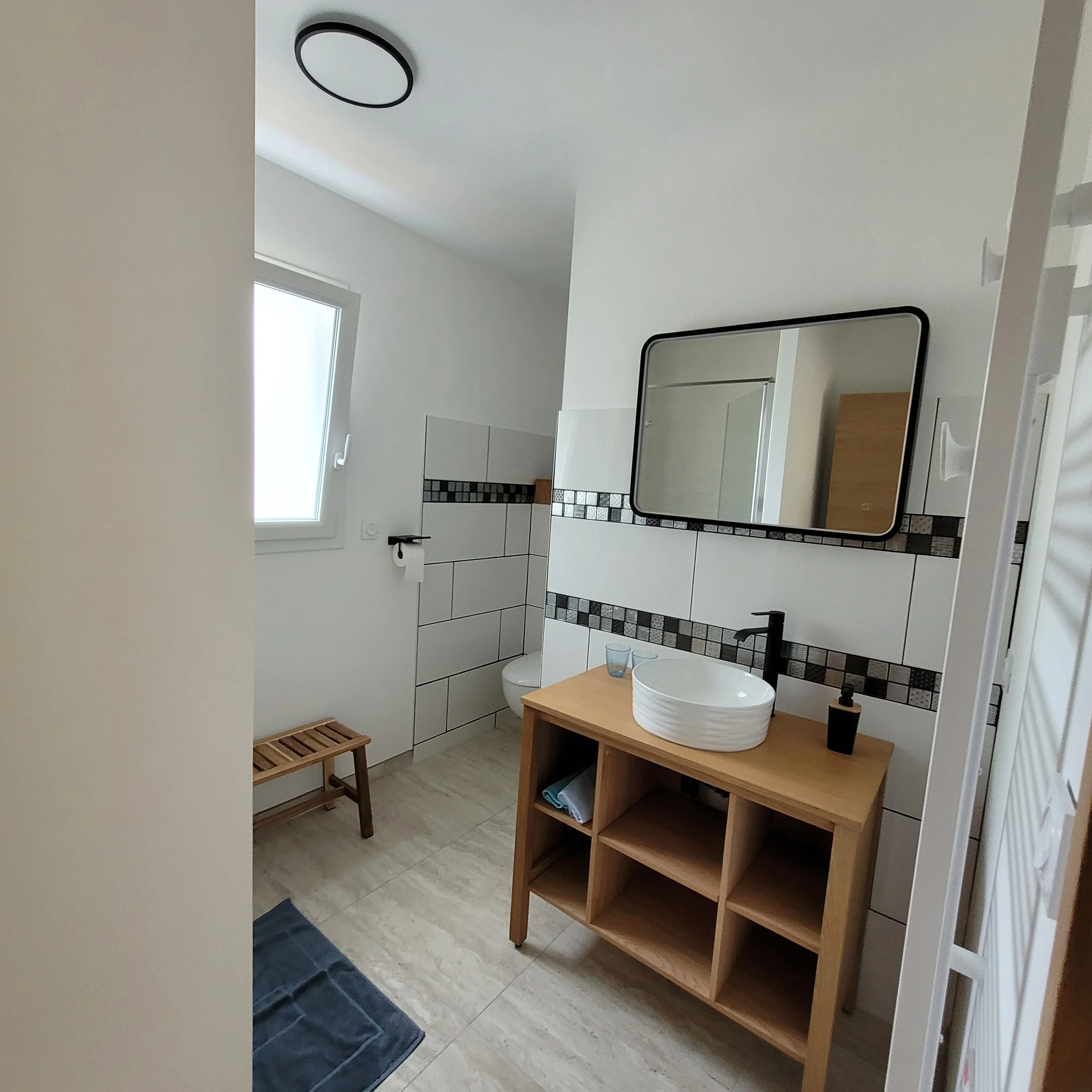 Une salle de bain moderne avec un lavabo rond blanc sur un meuble en bois, un miroir rectangulaire, un WC, un tapis de bain bleu foncé, une fenêtre laissant entrer de la lumière naturelle, et des carreaux noirs et gris en décoration derrière le lavabo.