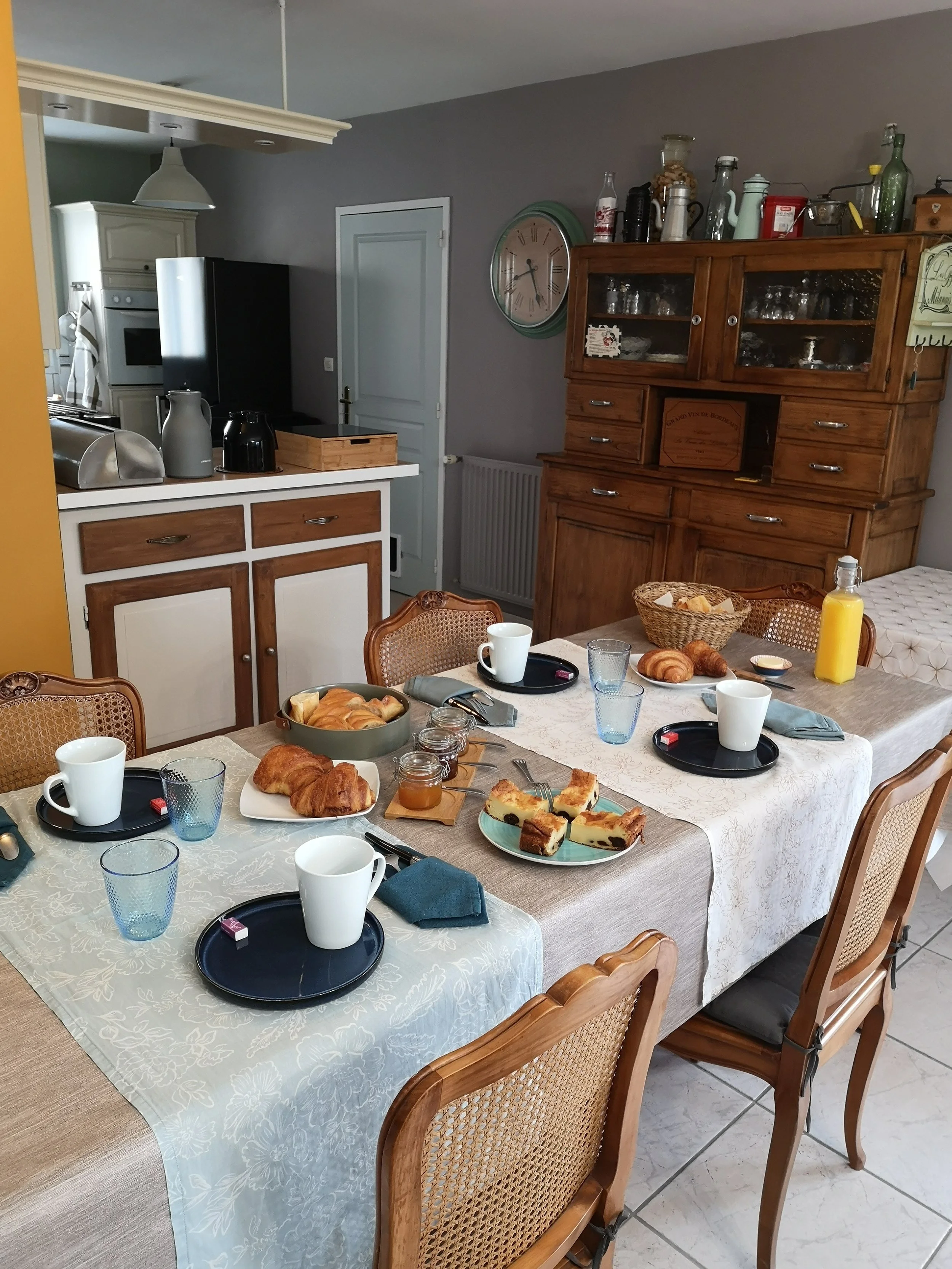 Une table de petit-déjeuner avec croissants, gâteaux et boissons, entourée de chaises en bois, dans une cuisine maison avec vaisselier en bois et horloge murale.