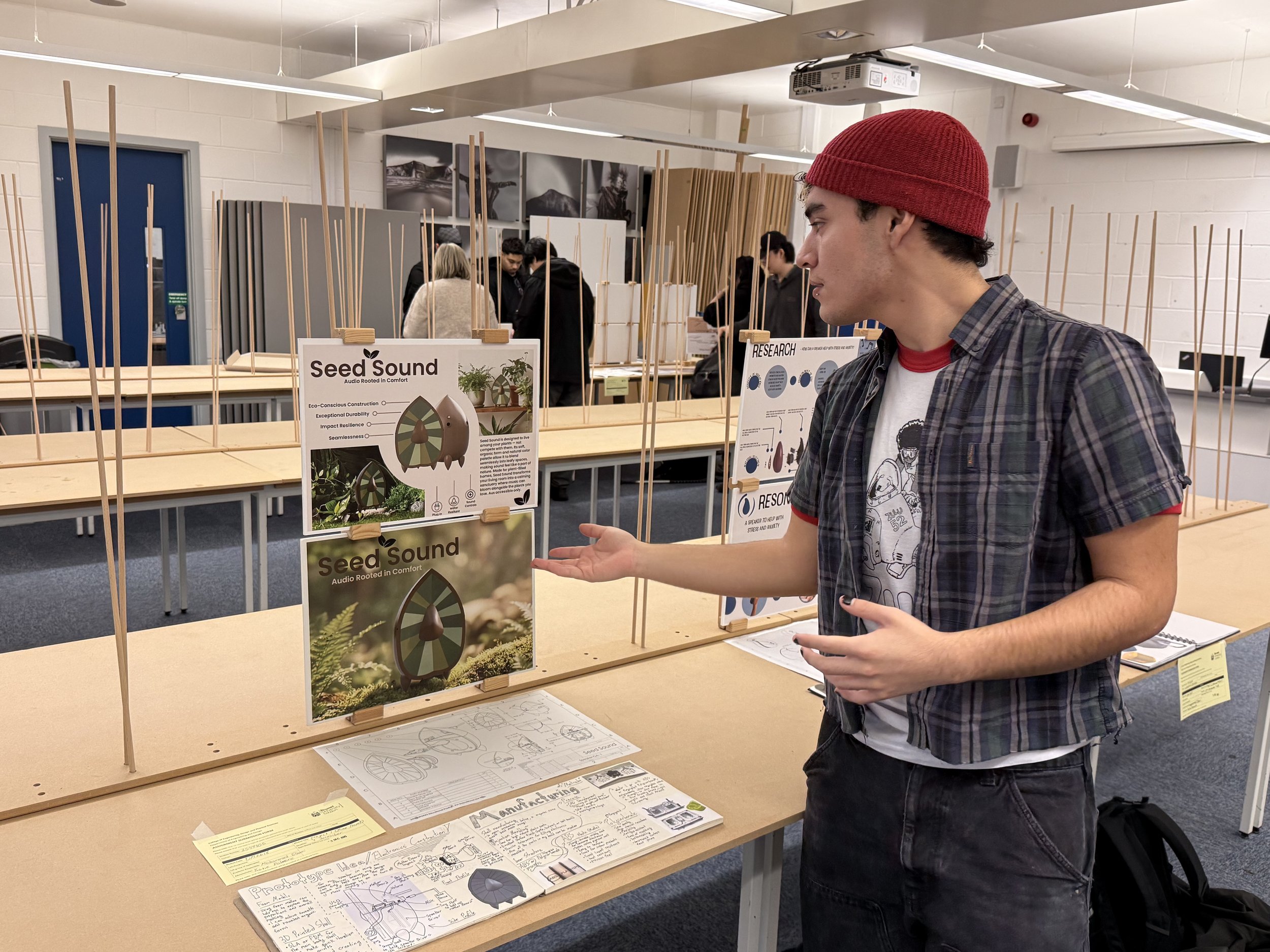 Young man in plaid shirt and red hat presenting a project at a science fair, with display boards about 'Seed Sound' and other research.