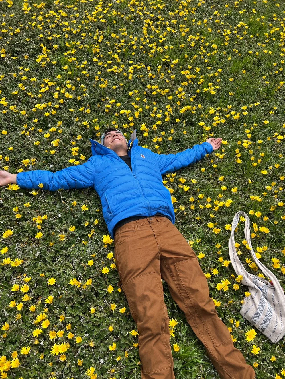 A person lying on a field of small yellow flowers with arms stretched out, smiling, wearing a bright blue jacket and brown pants, with a bag nearby.