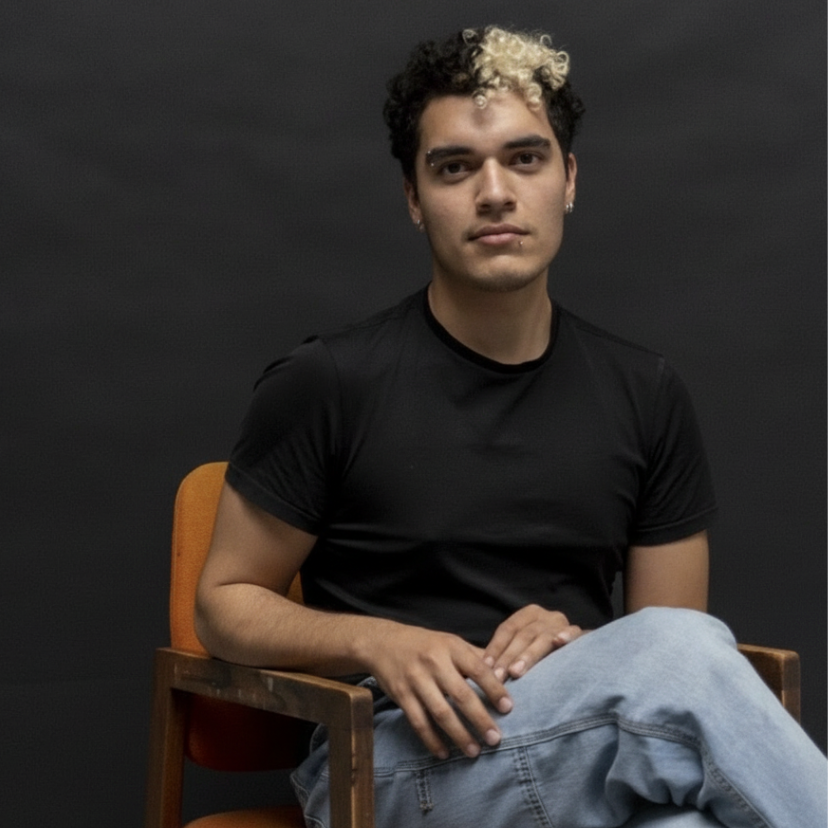Young man with curly two-toned hair, wearing a black t-shirt and light jeans, sitting on an orange chair against a dark background.