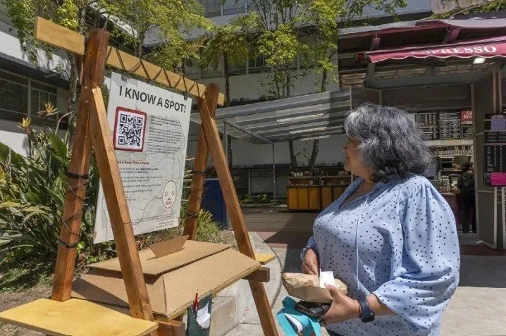 A woman with gray hair in a blue polka-dot blouse looking at an outdoor QR code sign with a woman behind a kiosk in the background.