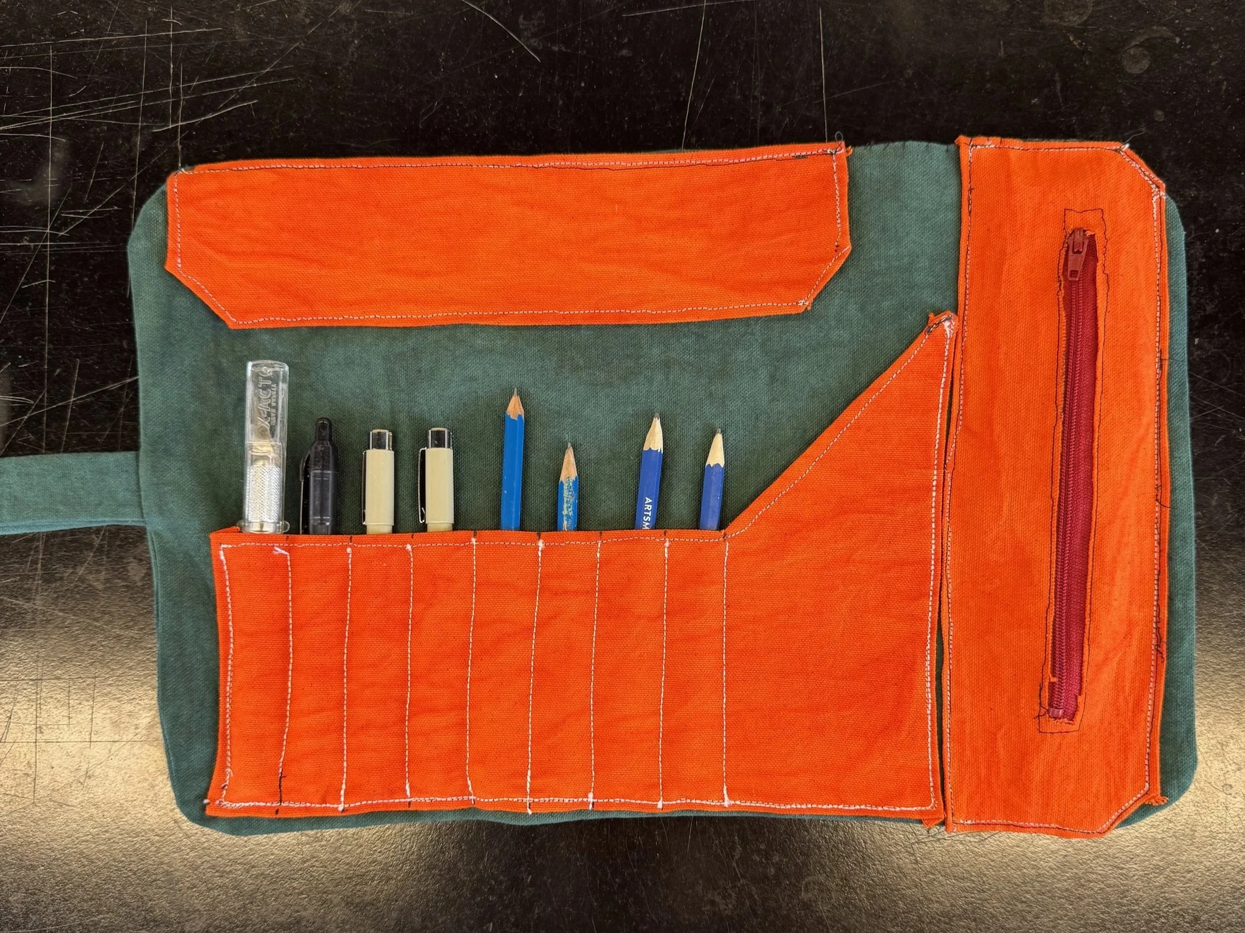 Open green and orange fabric pencil case with pens, pencils, small light, and a zipper pocket