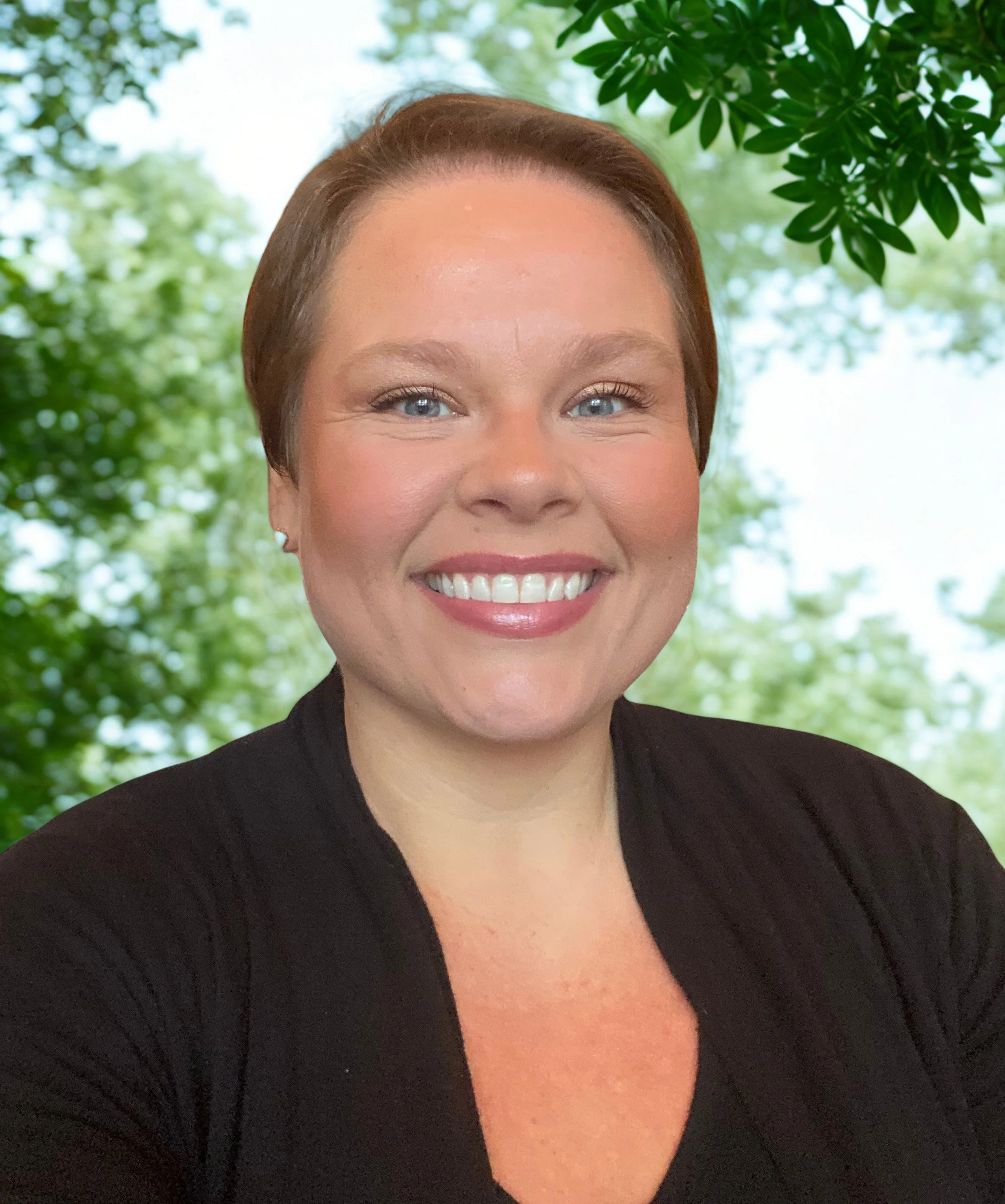 A woman with short brown hair, blue eyes, and a bright smile, wearing a black top, with a leafy green outdoor background.