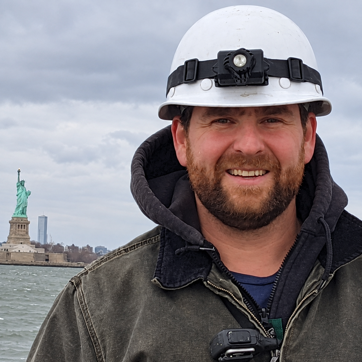 Peter Mogl wearing a hard hat with a headlamp, standing outdoors near a body of water, with the Statue of Liberty and New York City skyline in the background.