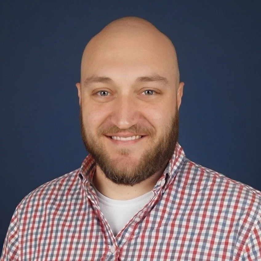 Headshot Michael De Jong, wearing a red and white checkered shirt against a dark blue background.