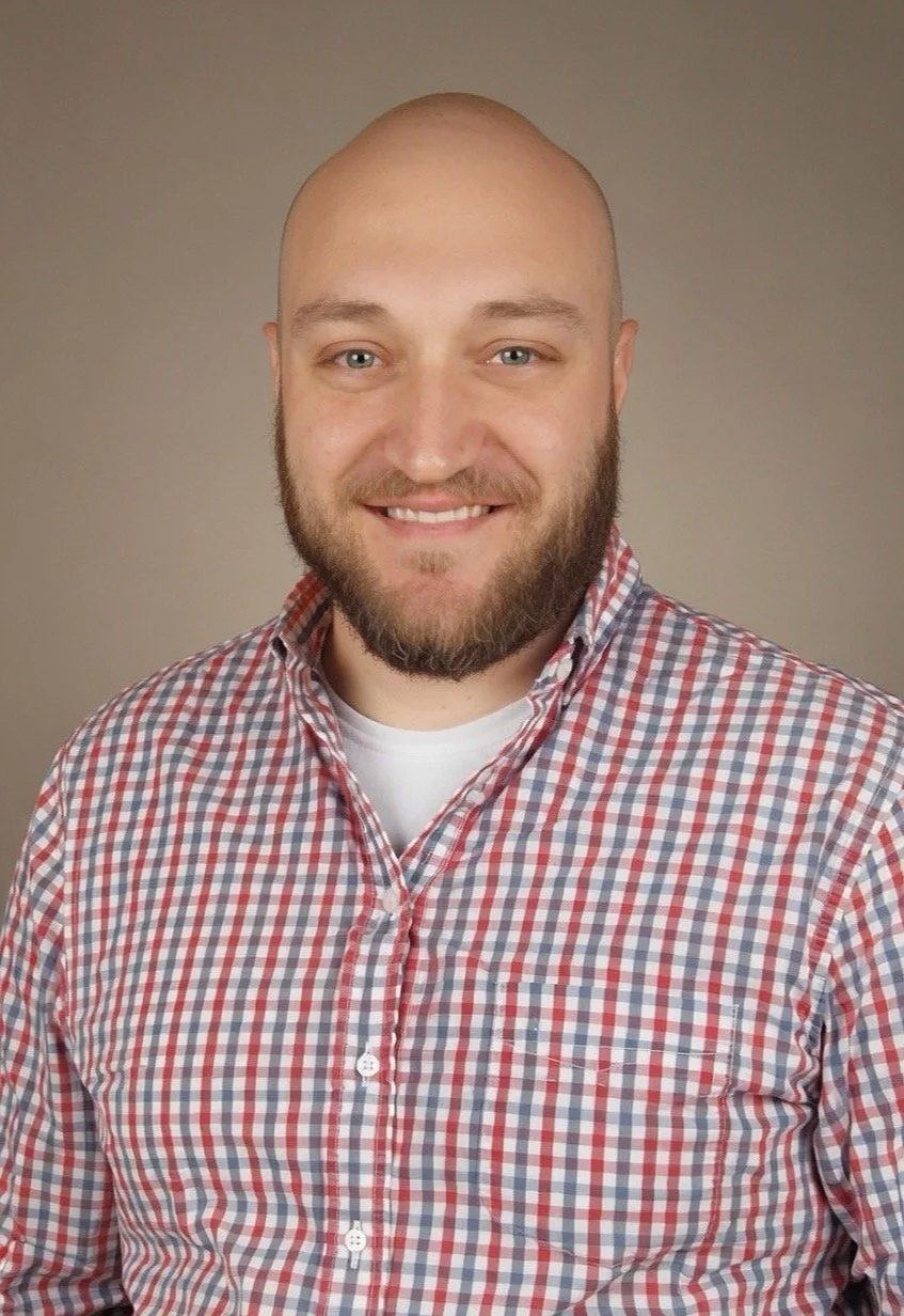 A man with a bald head and beard smiling, wearing a red, white, and blue checkered shirt over a white t-shirt, standing against a neutral background.