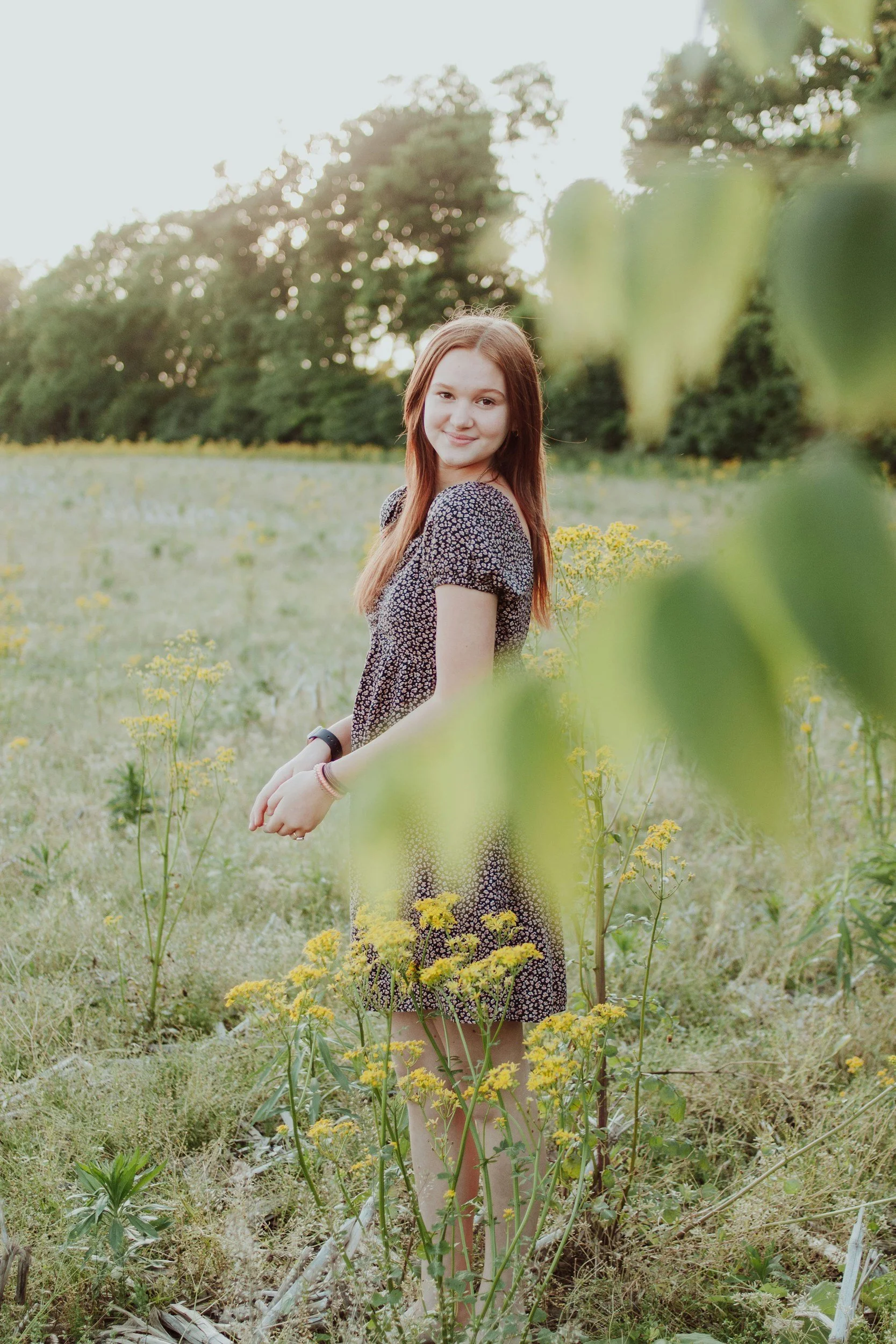 A young woman with long red hair standing in a grassy field with yellow flowers, smiling at the camera, with trees and sunlight in the background.