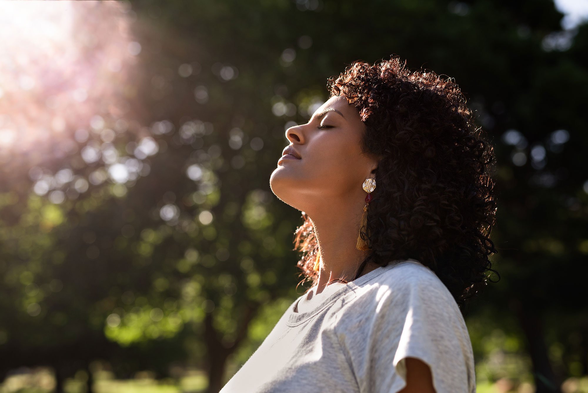 A woman with curly hair and earrings stands outdoors with her eyes closed and face tilted towards the sun, enjoying the sunlight and fresh air.