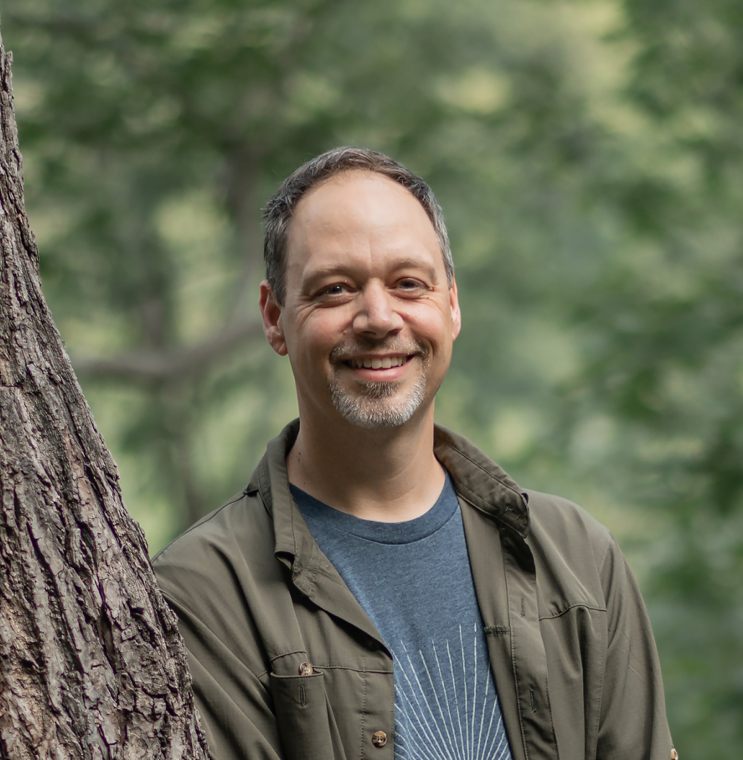 Jeff Gillespie smiling with short dark hair and a goatee stands outdoors in front of a tree and a blurred green forest background.