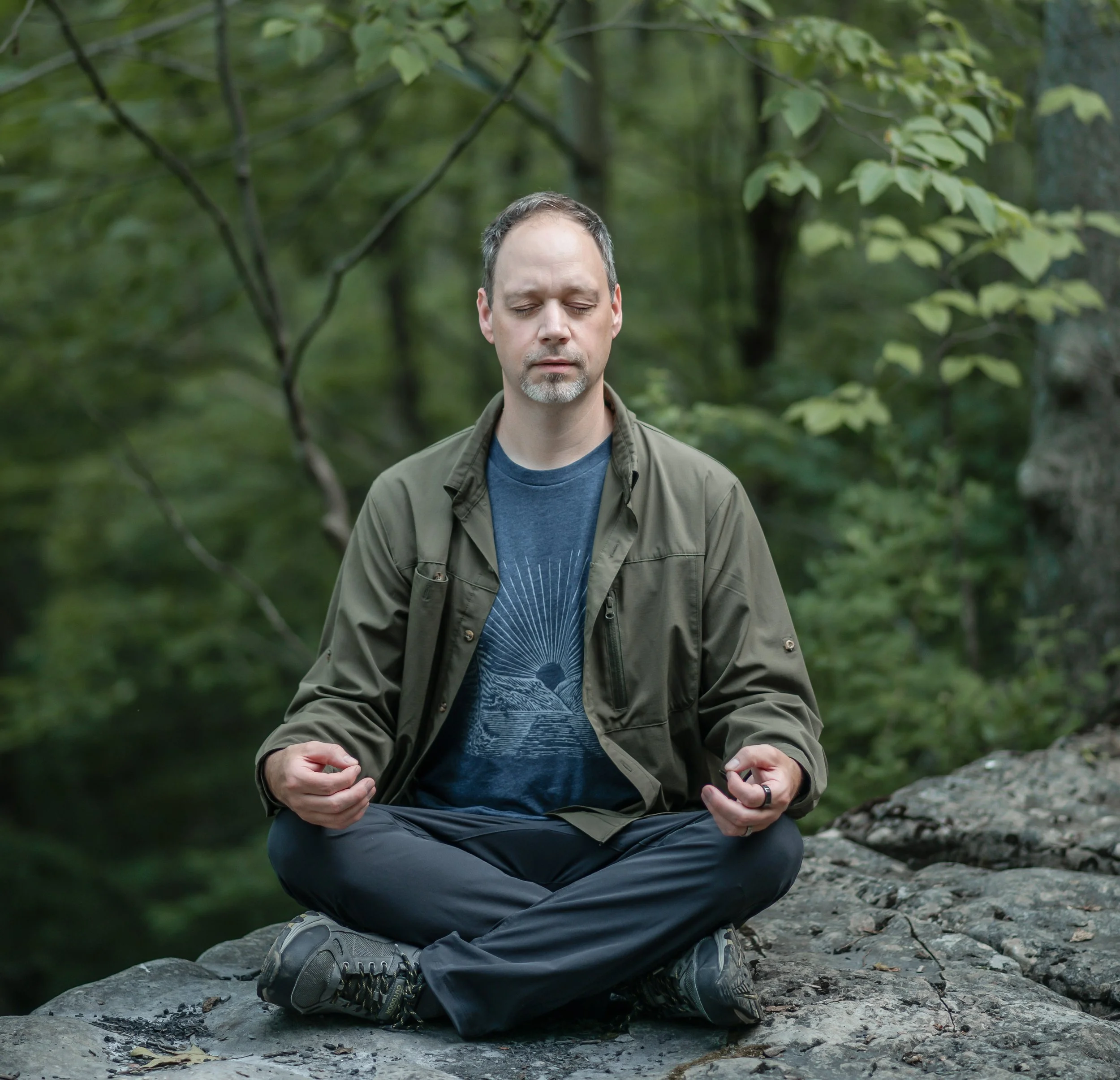 Jeff Gillespie sitting cross-legged on rocks in a forest, meditating with eyes closed.