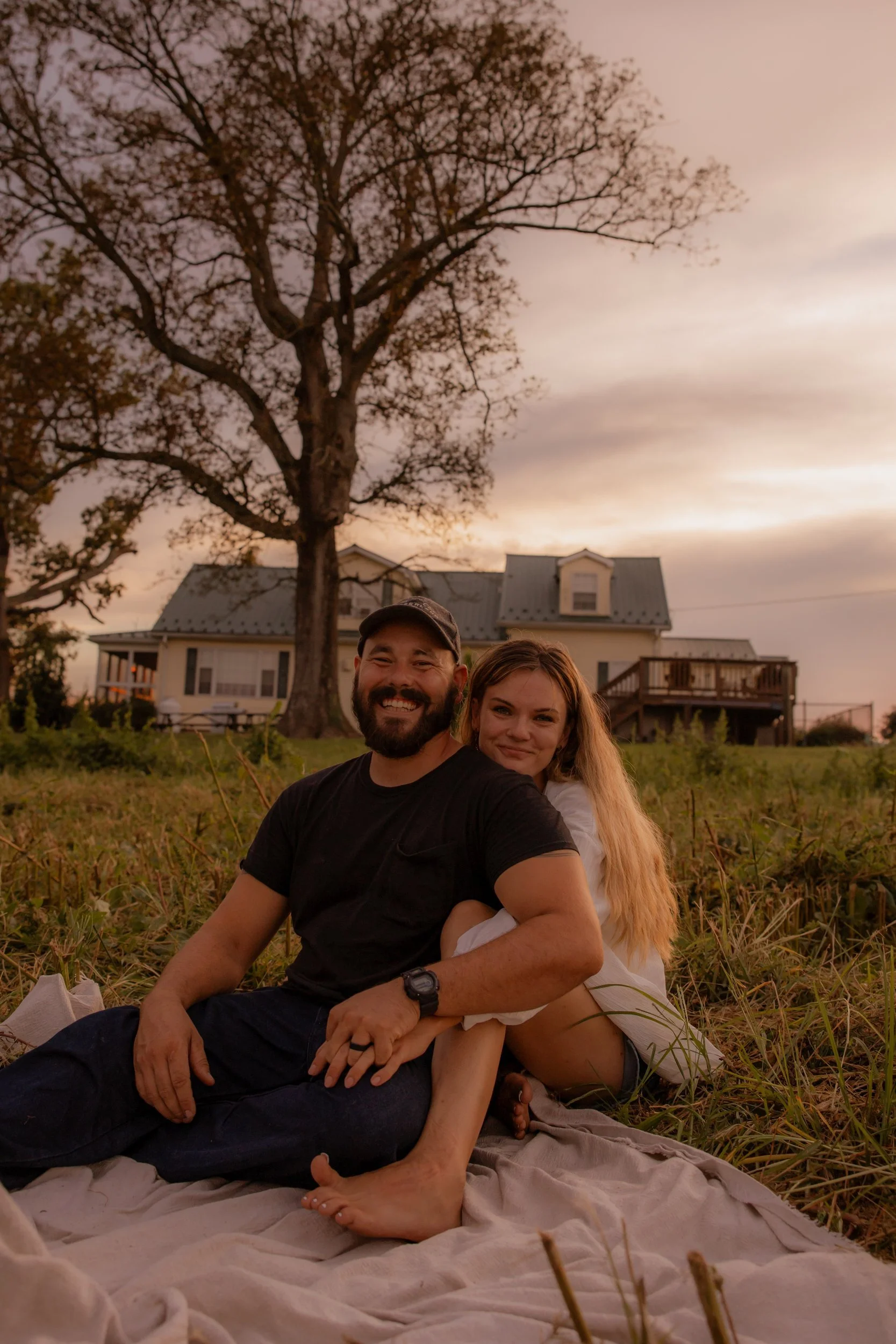 A smiling man and woman sitting closely together on a blanket outdoors during sunset, with a large tree and house in the background.