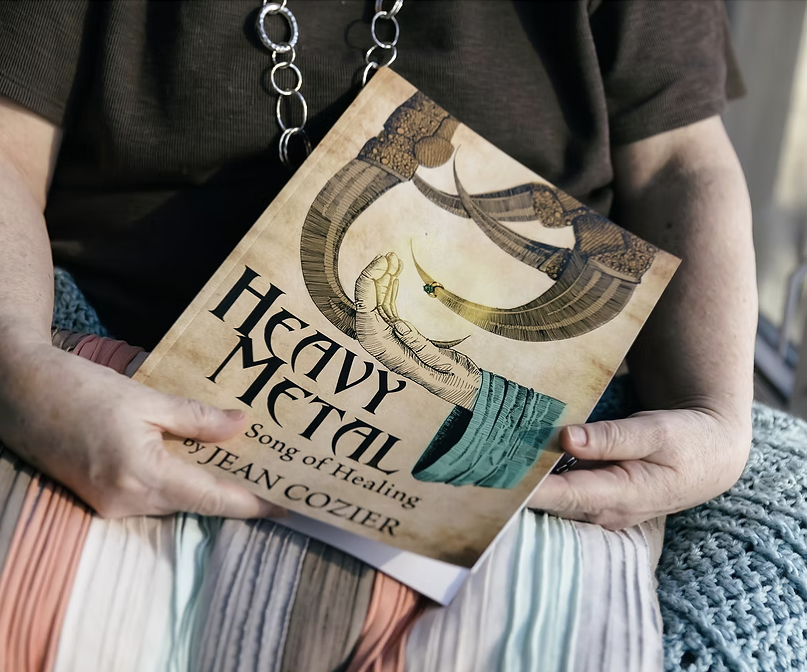 A person holding a book titled 'Heavy Metal: A Song of Healing' by Jean Cozier. The book cover features an illustration of a claw holding a ring.