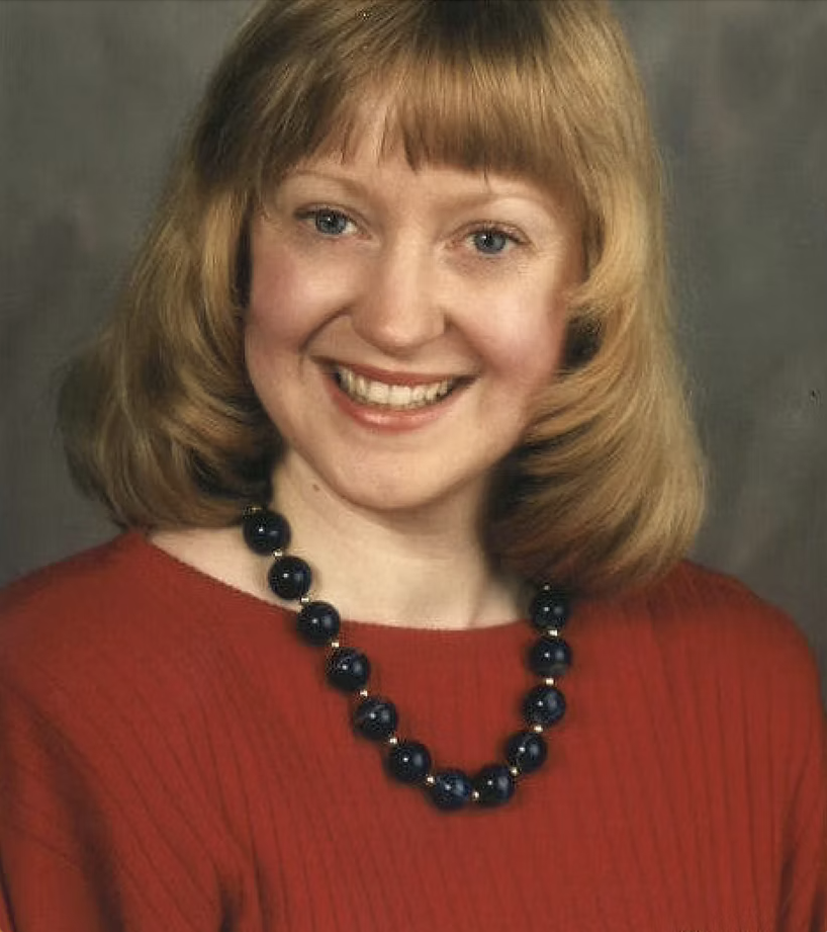 A woman with shoulder-length red hair and blue eyes smiling in a red top and black beaded necklace against a dark background.