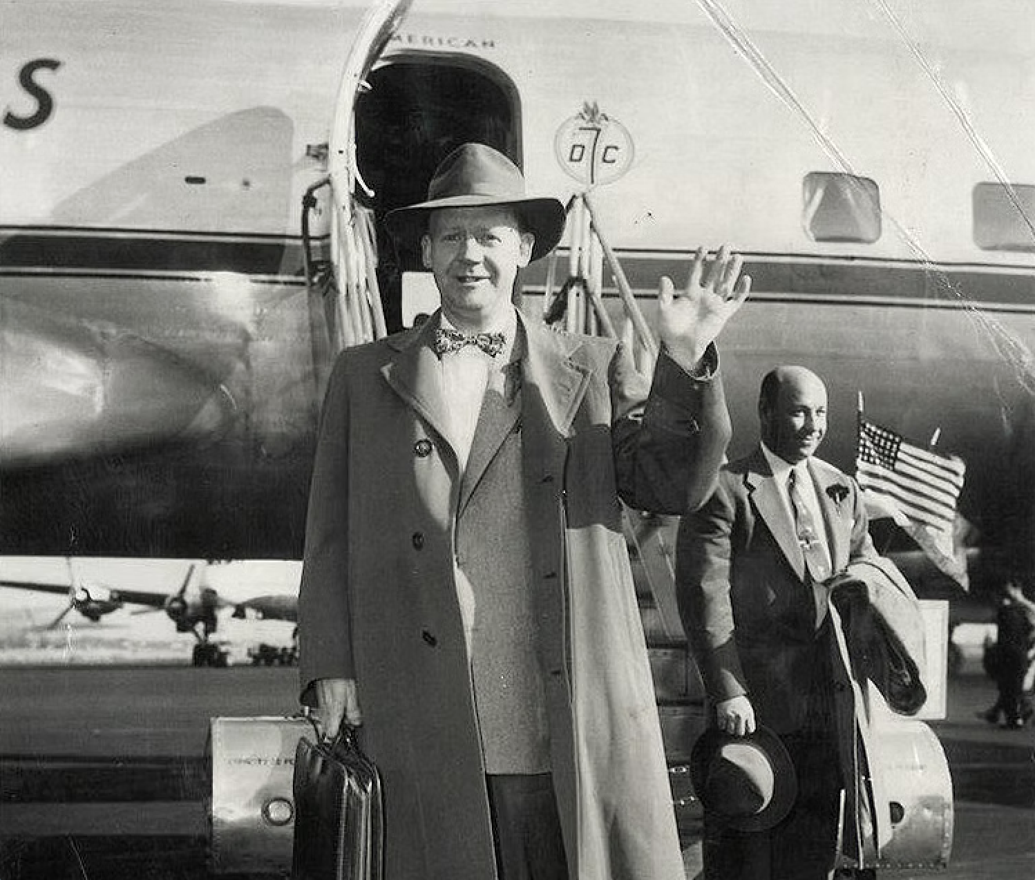 Black and white photo of two men arriving at an airport, one is waving, the other is holding a hat and a suitcase, with a plane and American flag visible in the background.