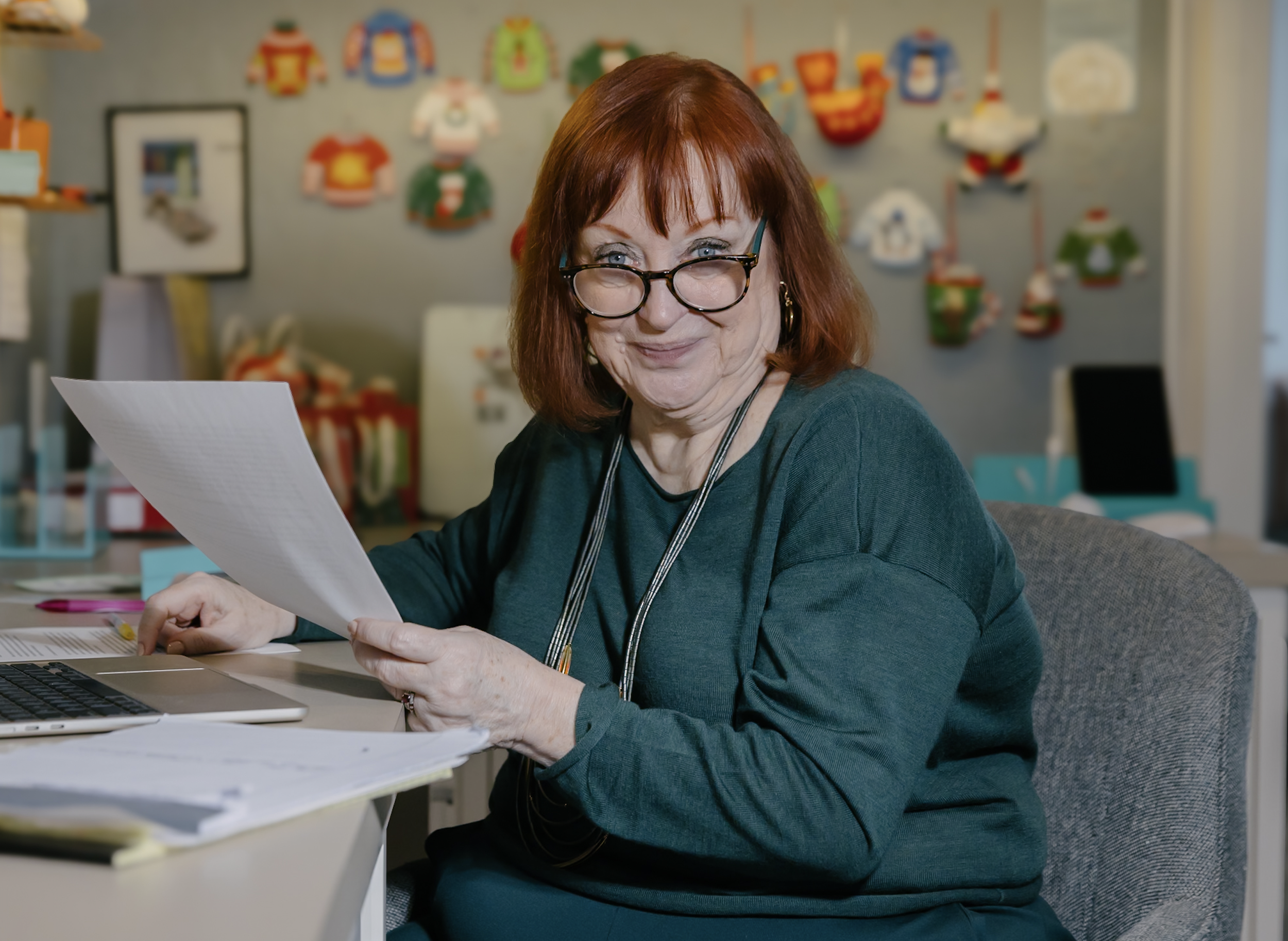 A woman with red hair and glasses smiling while looking at a piece of paper at a desk in an office decorated with colorful holiday-themed artwork.