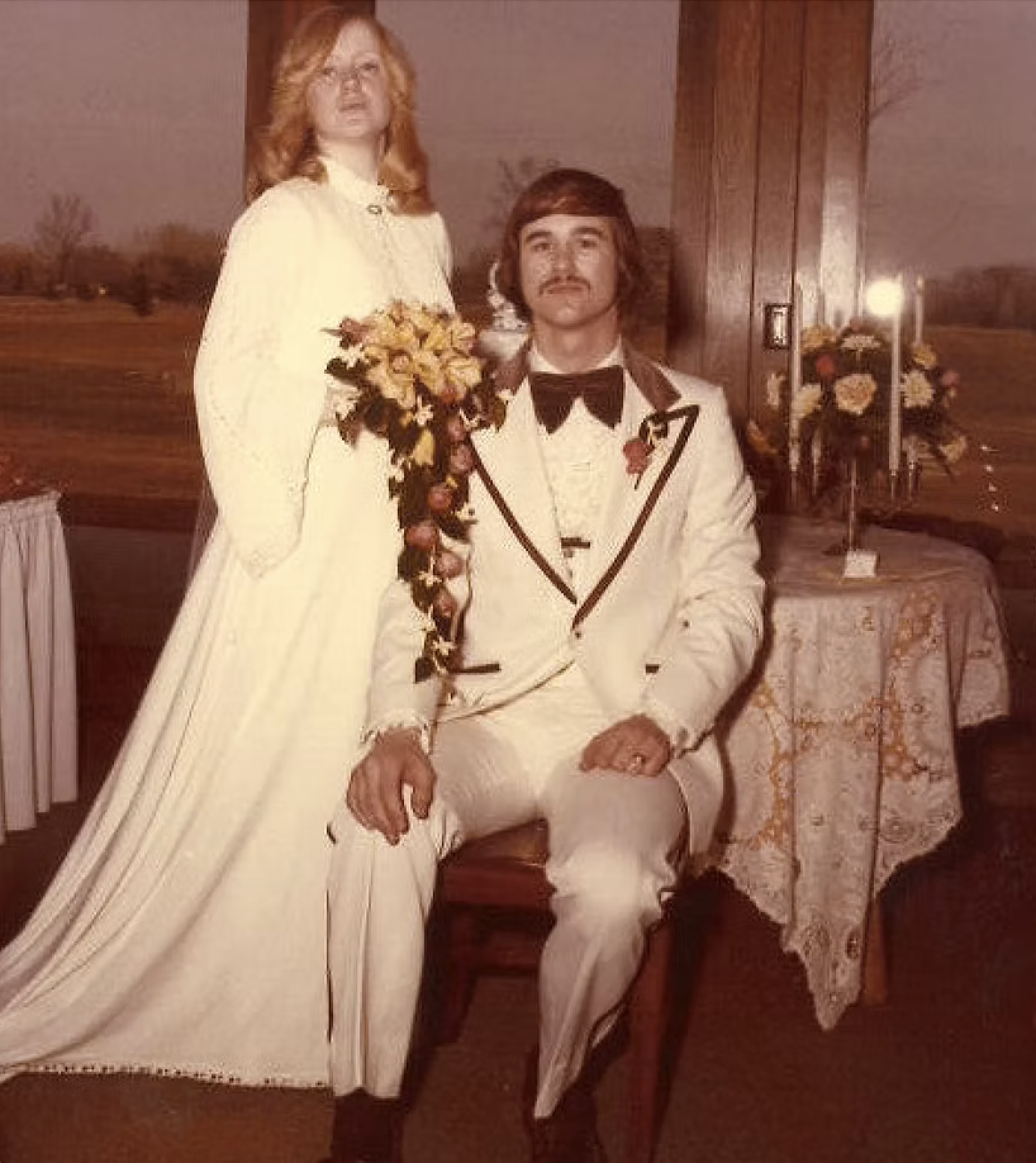 A couple dressed in vintage wedding attire, with the woman holding a bouquet of flowers, posing indoors with a rural landscape visible through a window in the background.
