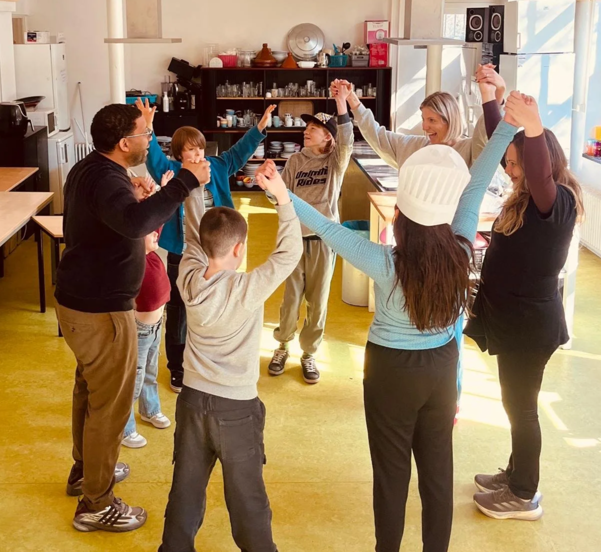 Group of children and adults holding hands in a circle, playing a game or dancing in a kitchen or community space, with sunlight streaming through windows.
