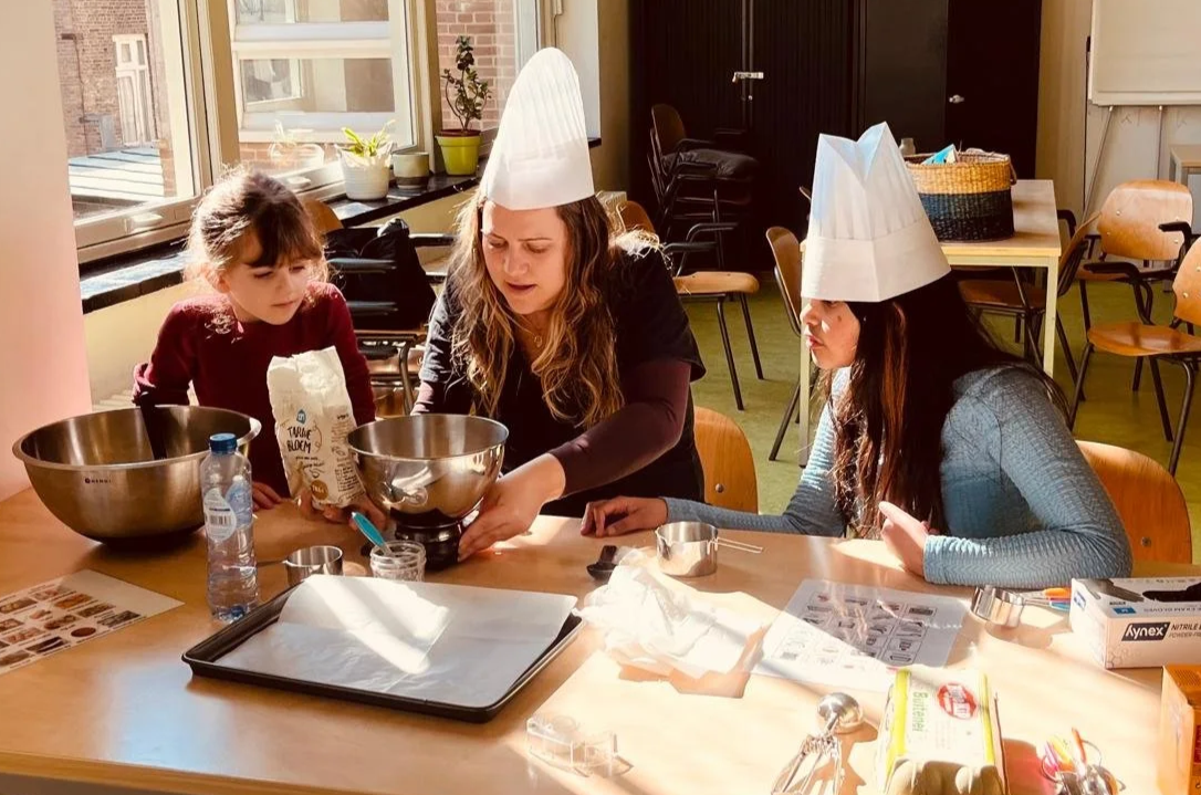 Three women, two young girls and one adult, wearing chef hats and aprons, engaged in a baking activity at a table with baking supplies in a sunlit room.